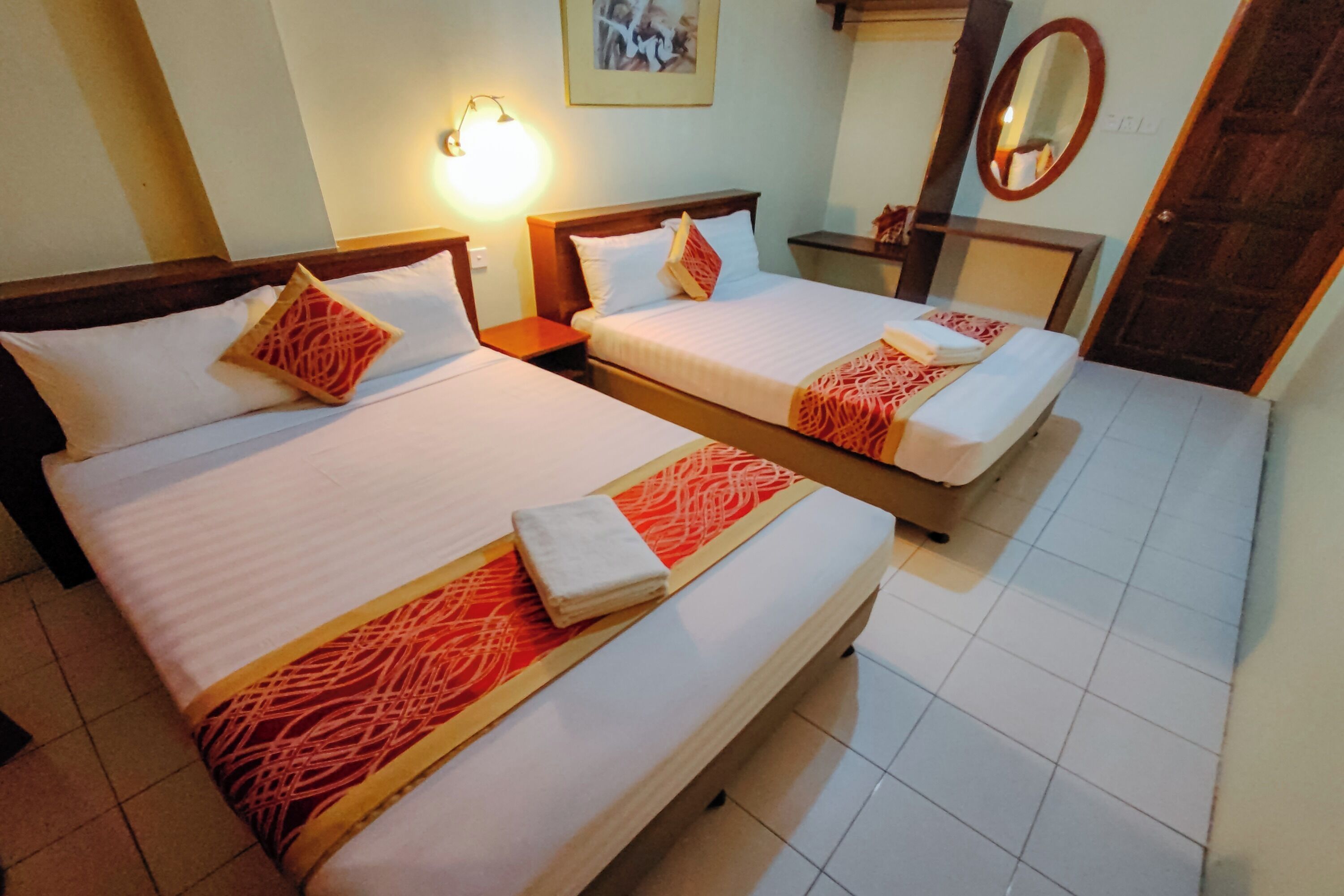 Habitación de hotel con dos camas, cada una con cabecero de madera, sábanas blancas y cubrelechos rojos estampados. Entre las camas hay una mesita de noche con una lámpara, y un espejo en la pared.