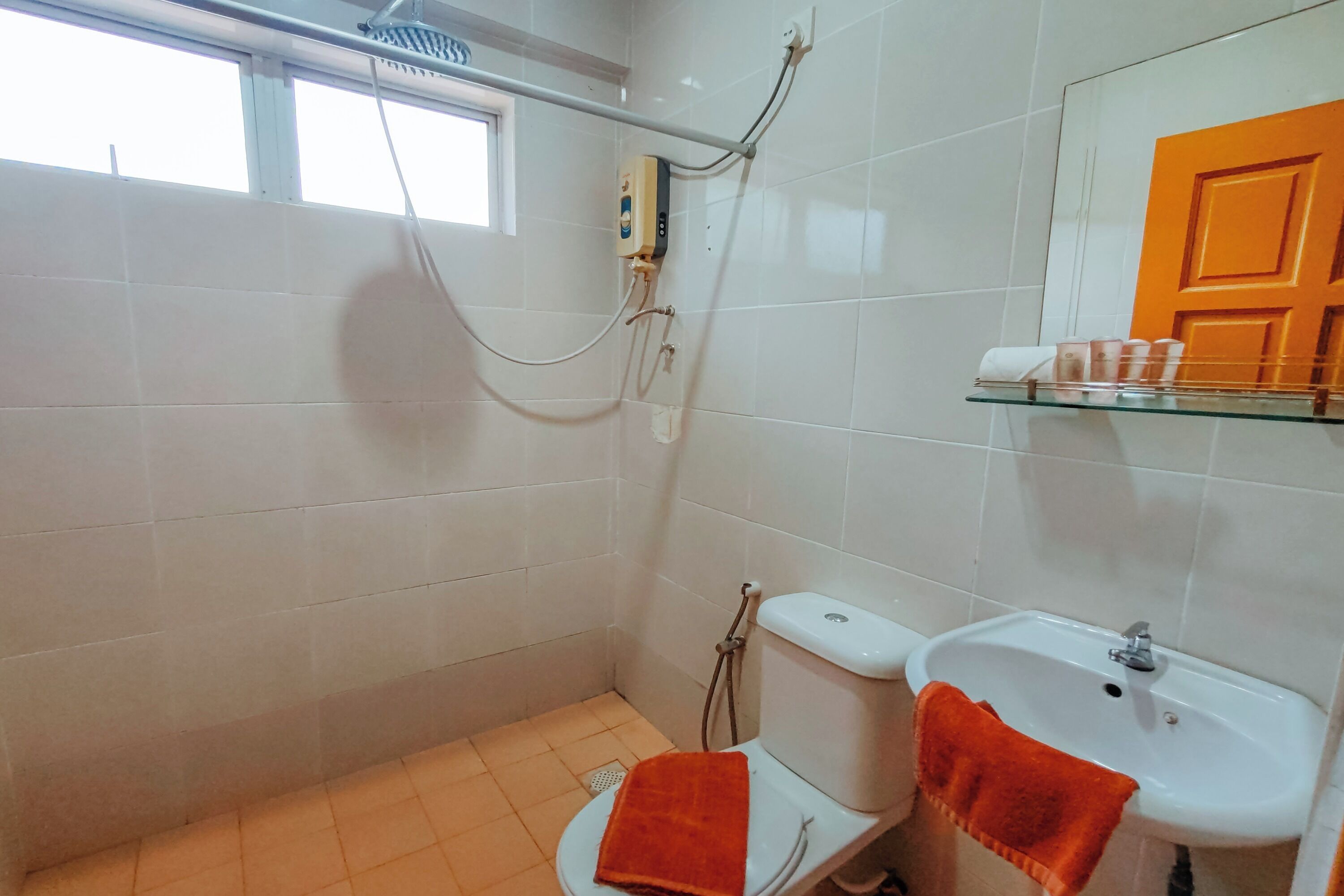 A bathroom with a toilet, sink, and shower. There is a window above the toilet, a towel hanging on the sink, and a shelf with toiletries.