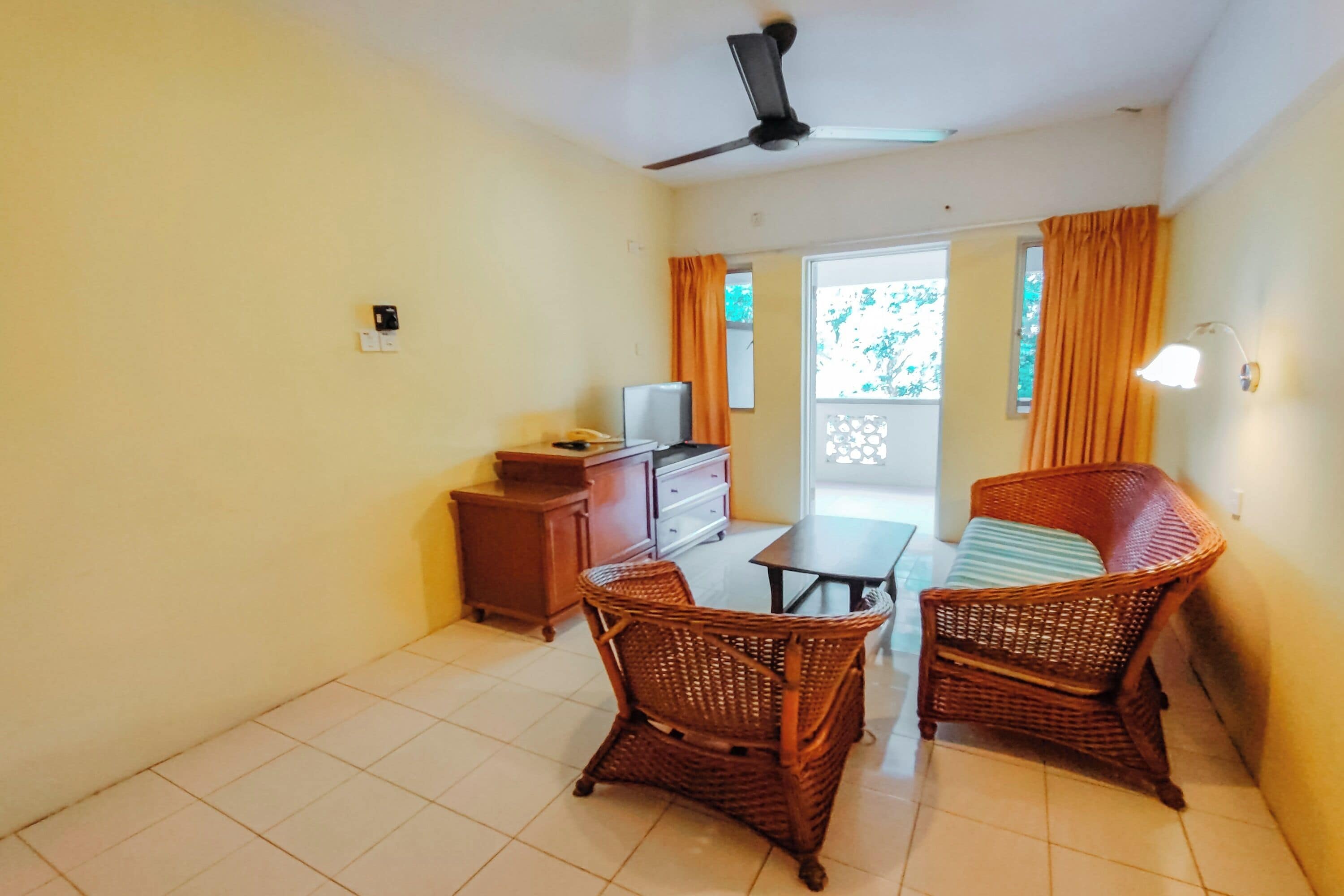 A room with a ceiling fan, a television, a wooden cabinet, wicker chairs, a table, and a view through a glass door.