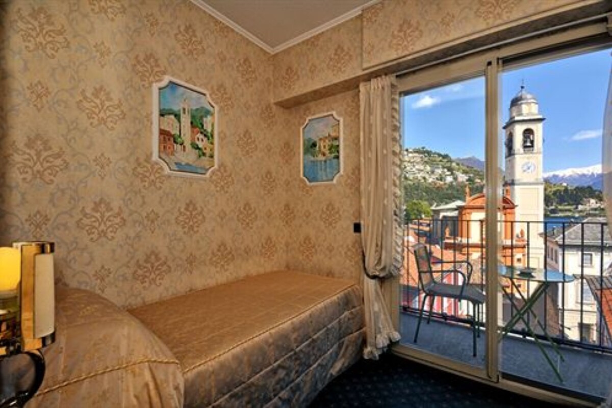 A room with a bed, a window with a view of a bell tower and mountains, and a balcony with chairs.