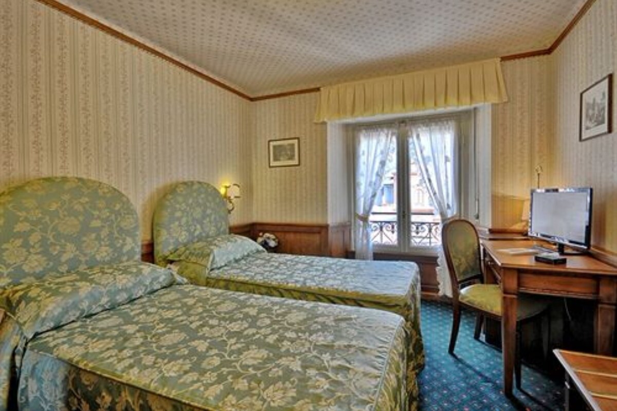 A hotel room with two single beds, a desk with a computer, a chair, and a window with curtains.