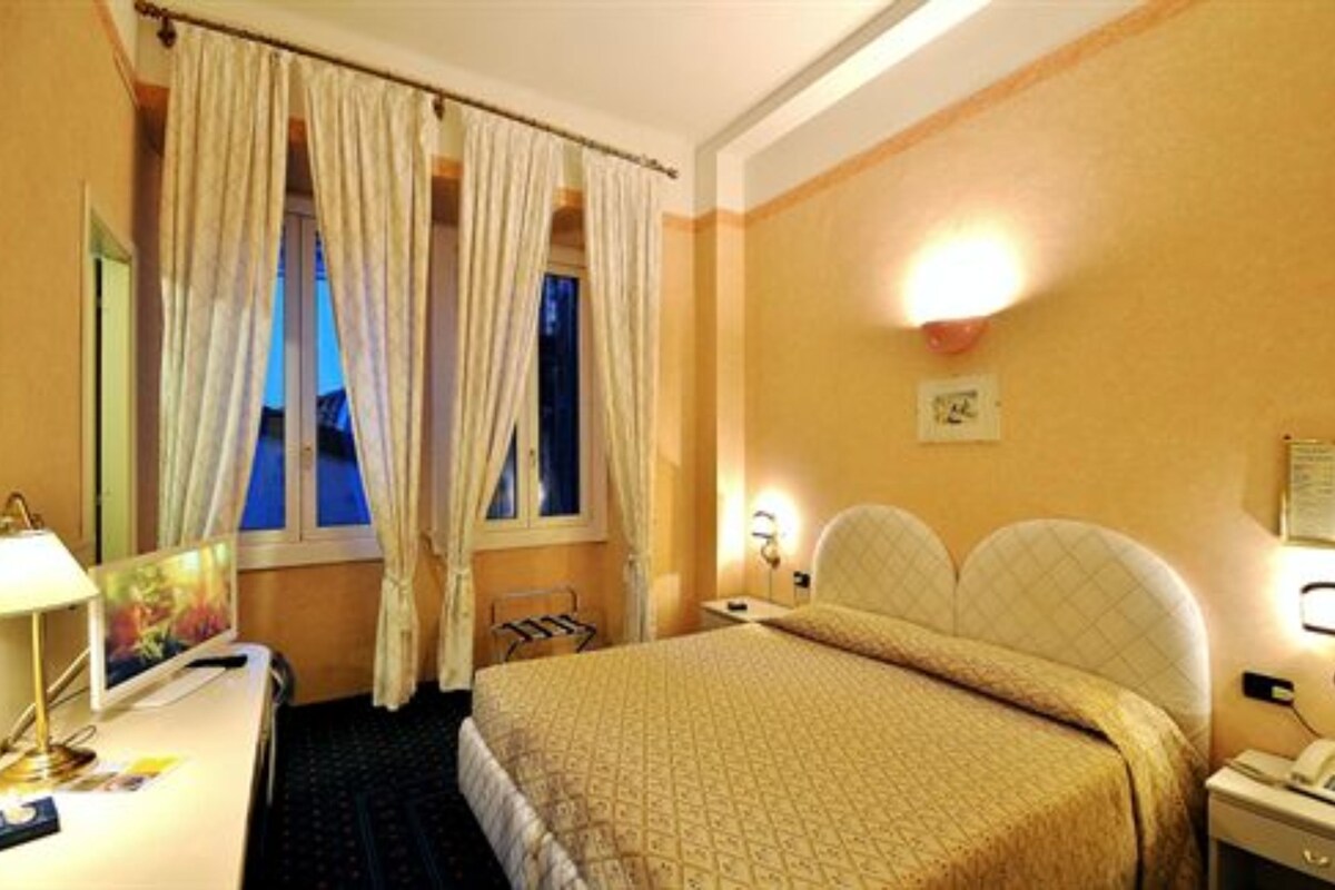 A hotel room with a bed, a desk with a lamp, a chair, and a window with curtains.