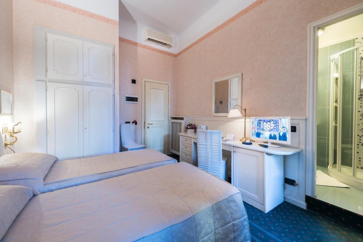 A hotel room with two beds, a desk with a computer and lamp, a mirror, and a shower area.