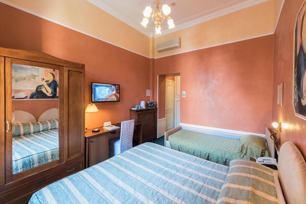 A hotel room with a large bed, a desk with a chair, a television, and a wardrobe.