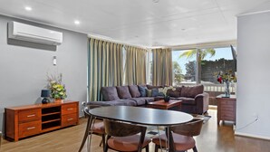 A modern living room with a sectional sofa, a dining table with chairs, a wooden cabinet, and a large window with curtains.