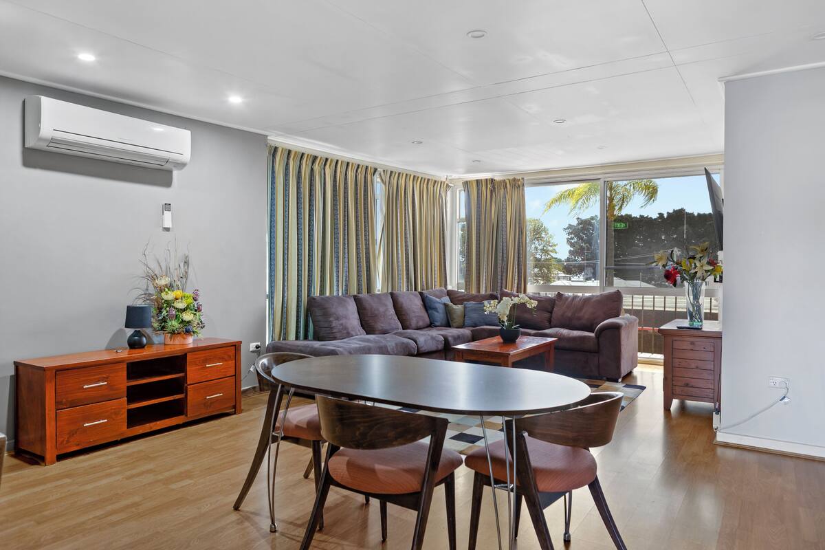 A modern living room with a sectional sofa, a dining table with chairs, a wooden cabinet, and a large window with curtains.