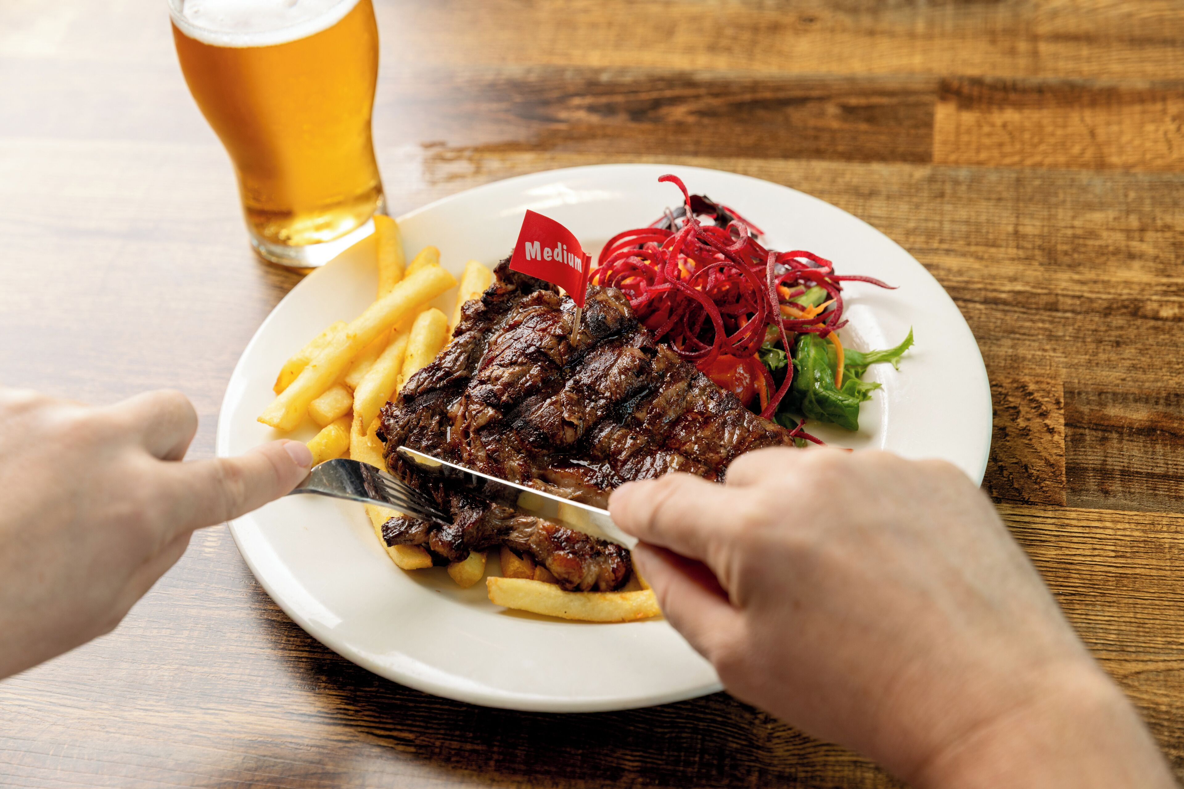 A plate with steak, fries, and a salad, and a glass of beer on a wooden table.