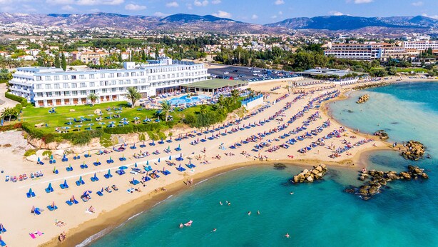 Un hôtel situé sur la côte, doté d’une grande piscine, d’une plage avec des chaises longues et offrant une vue sur la ville et les montagnes en arrière-plan.