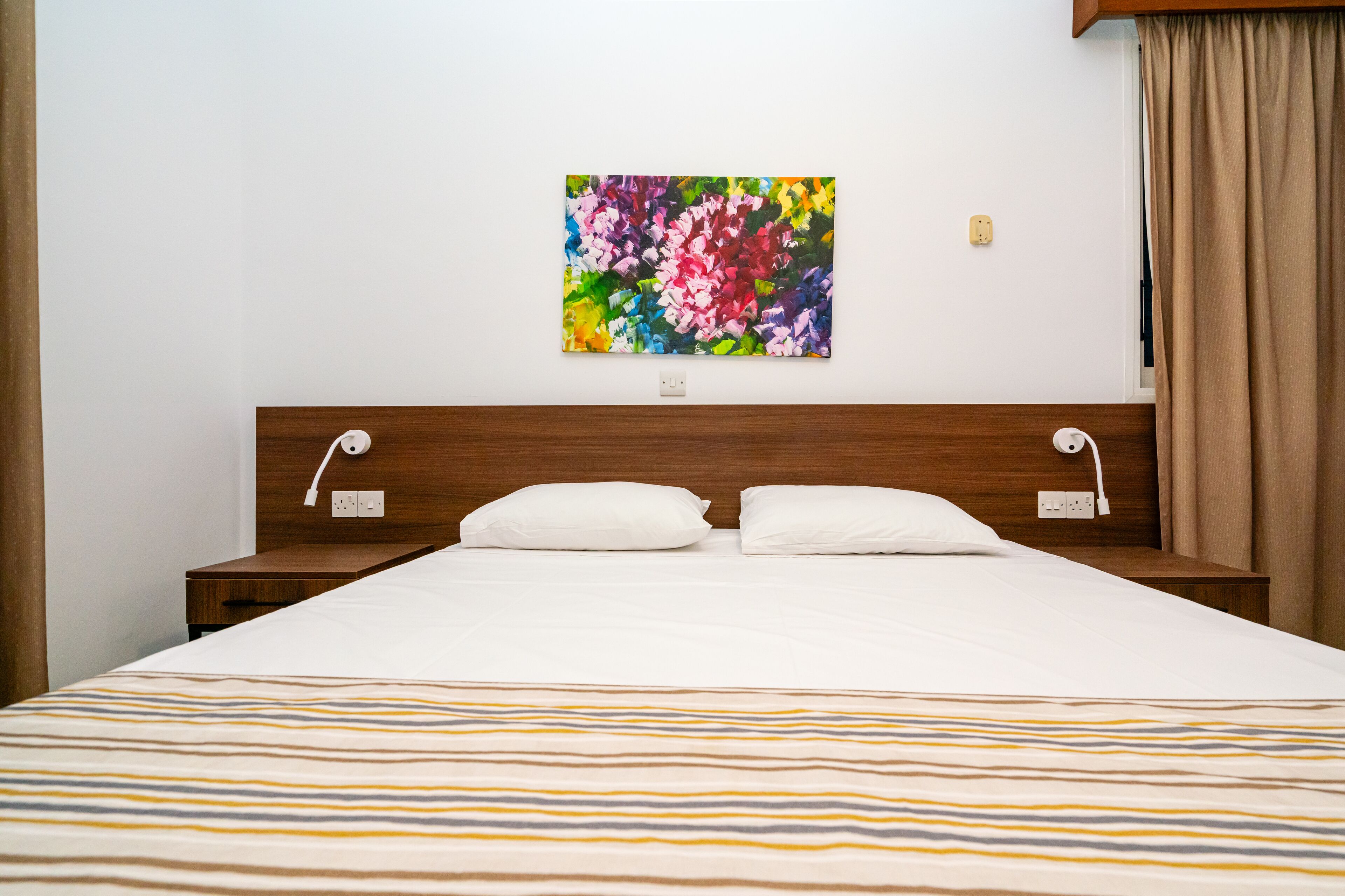 A neatly made bed with a striped bedspread, two white pillows, and a colorful abstract painting on the wall above.