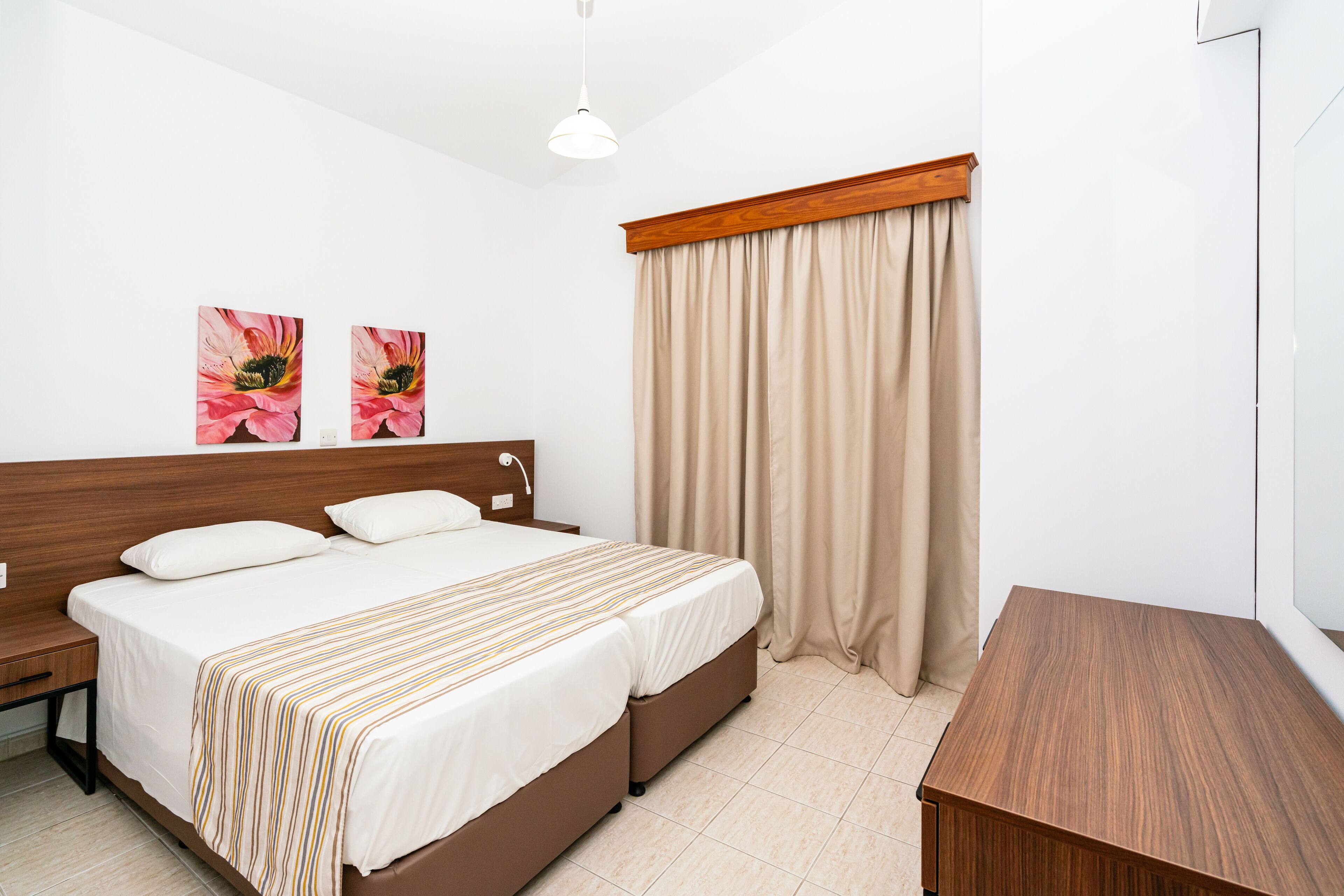 Ett välordnat hotellrum med en säng, sängbord, ett litet träbord, ett mindre träbord och ett fönster med beige gardiner.