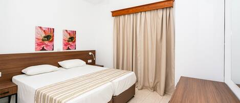 A neatly arranged hotel room with a bed, bedside tables, a wooden nightstand, a small wooden table, and a window with beige curtains.