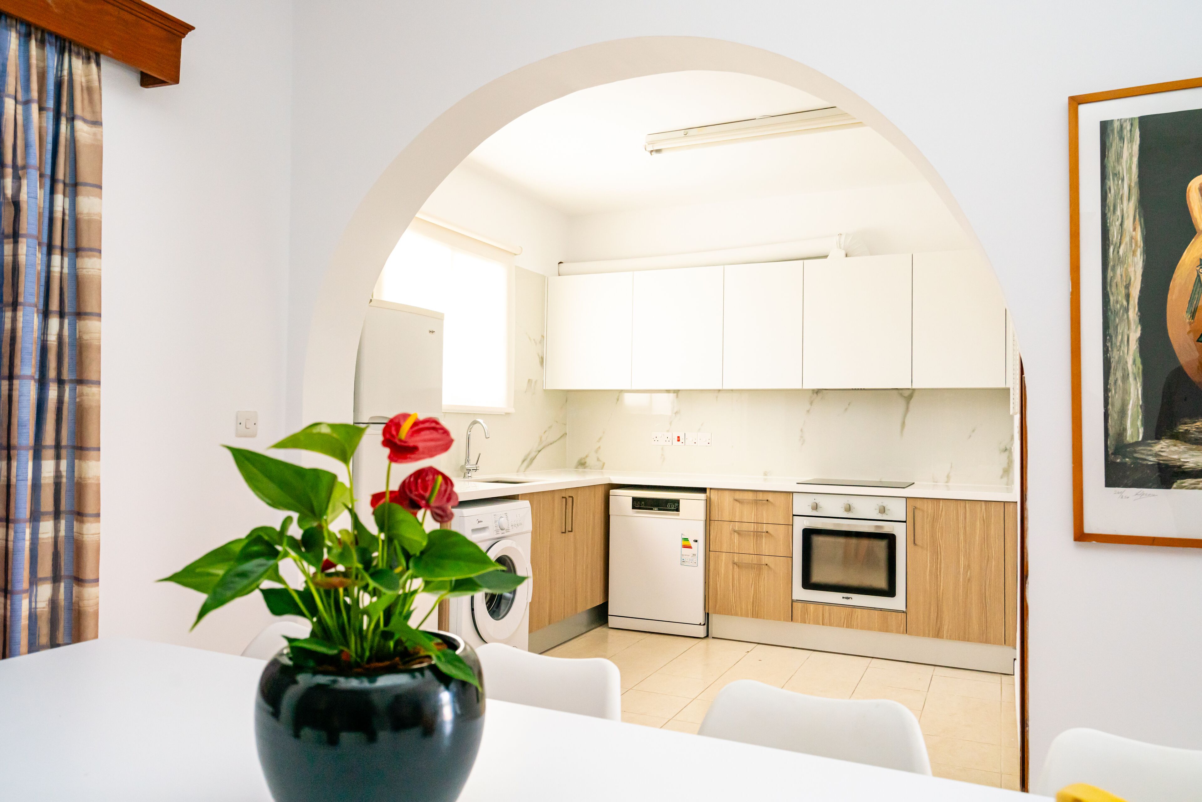 A modern kitchen with white cabinets, a dishwasher, and an oven. There is a dining area with a table and chairs, and a vase with flowers on the table.