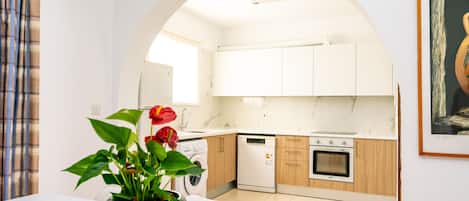A modern kitchen with white cabinets, a dishwasher, and an oven. There is a dining area with a table and chairs, and a vase with flowers on the table.