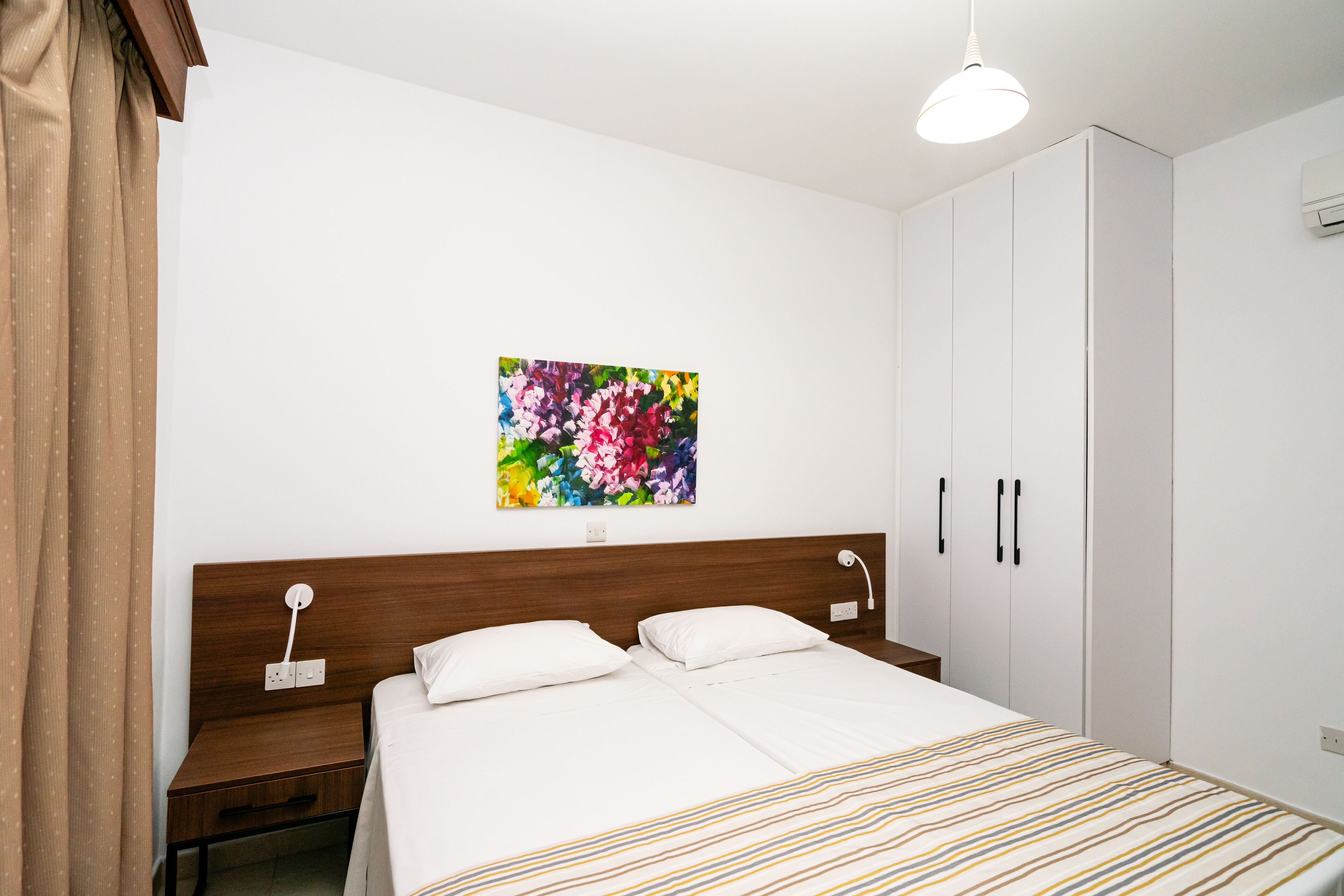 A bedroom with a bed, nightstands, a wooden headboard, a white wardrobe, and a colorful floral painting on the wall.