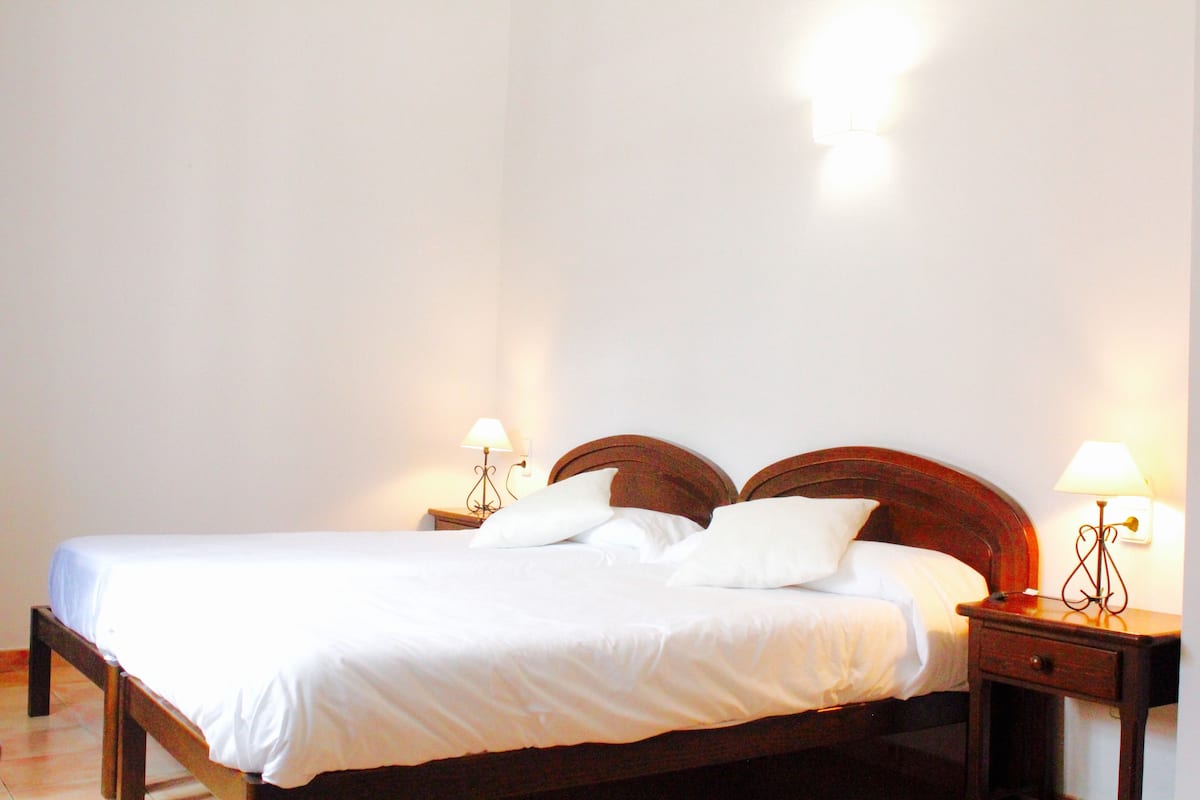 Una cama bien hecha con sábanas blancas, dos mesitas de noche con lámparas y un cabecero de madera.