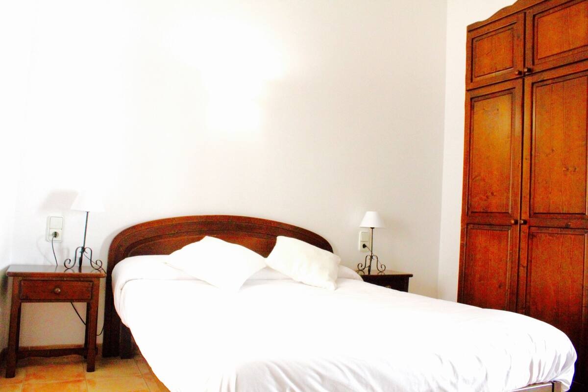 Una cama bien hecha con ropa blanca, dos mesitas de noche con lámparas y un armario de madera.