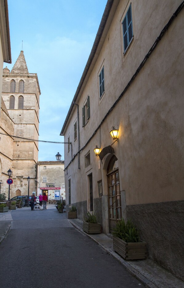 A narrow street with historic buildings, including a tower with arched windows and a building with a wooden door and a small garden.