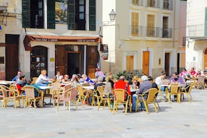 An outdoor cafe with wicker chairs and tables, a restaurant with a brown awning, and a building with balconies and shutters. - Hotel Can Font (Sineu)