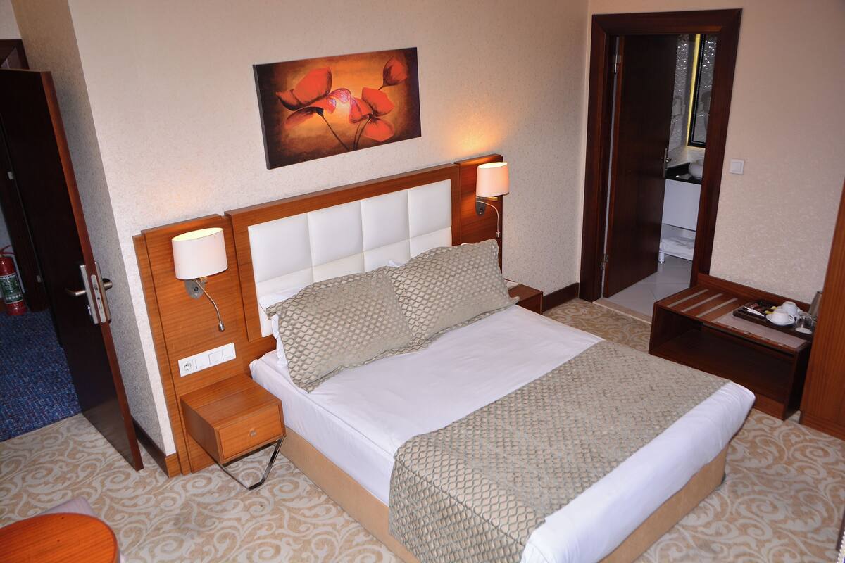 A hotel room with a bed, bedside lamps, a nightstand, a small table, a mirror, and a painting on the wall.