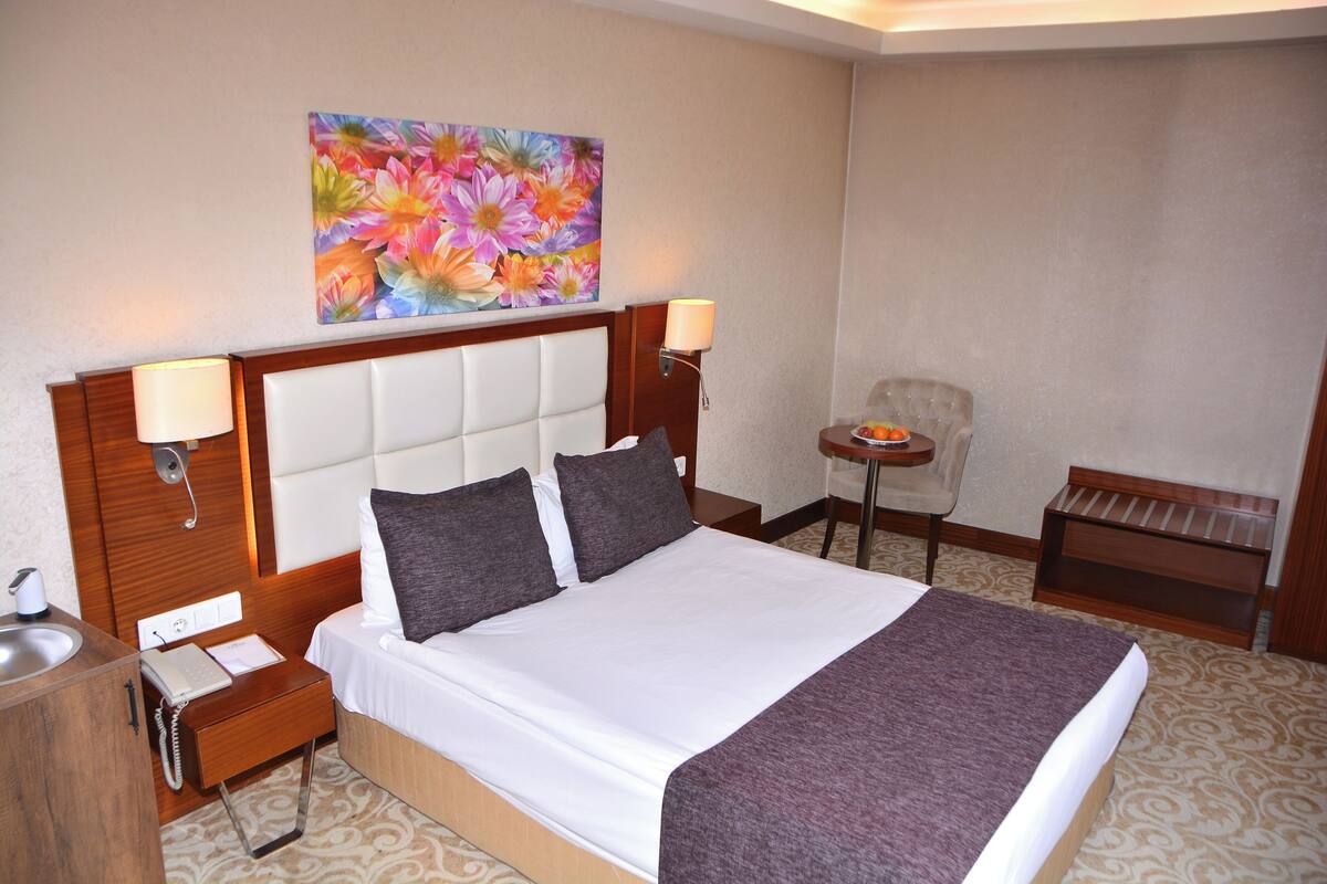 A hotel room with a large bed, two bedside lamps, a small table with a chair, and a painting on the wall.