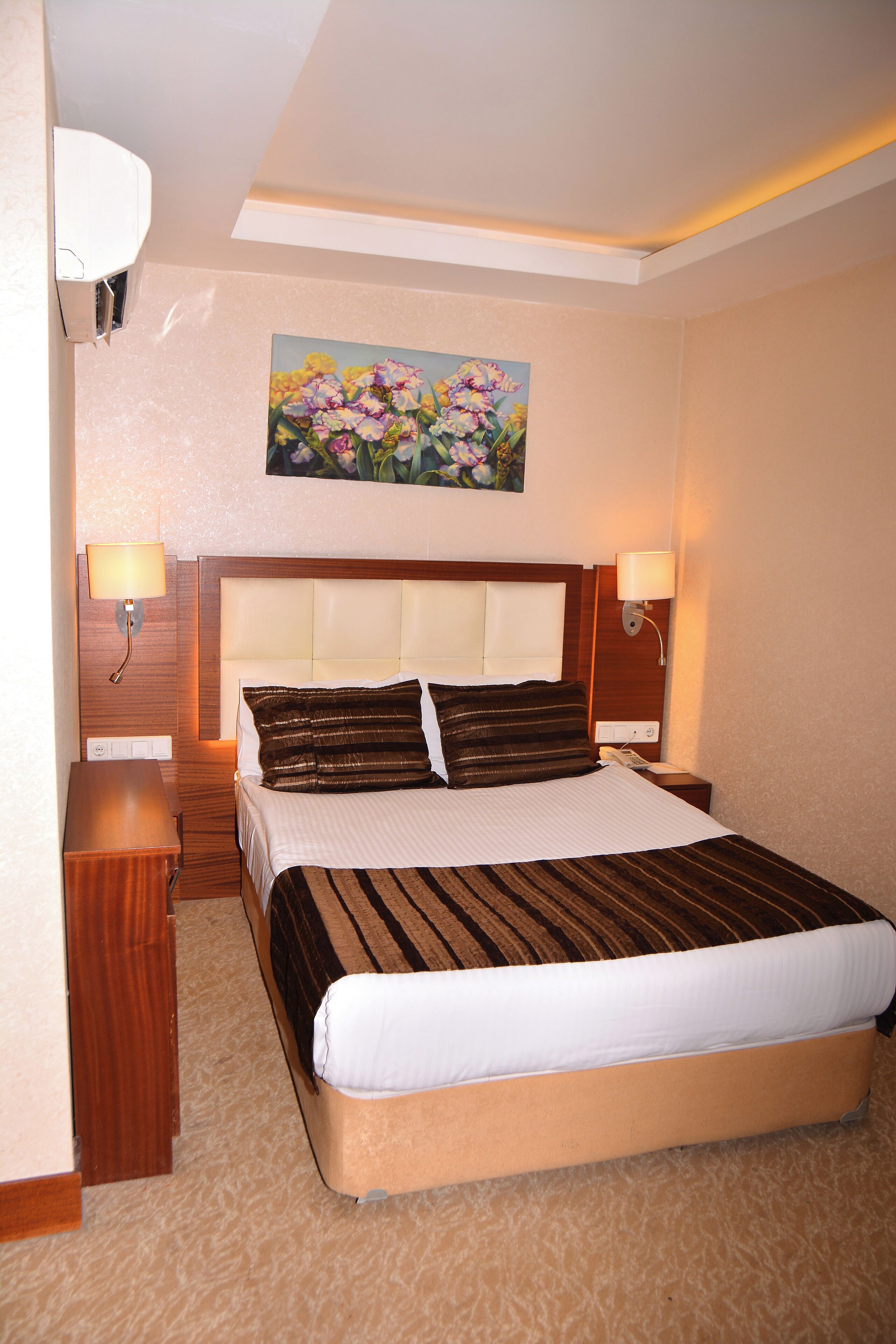 A hotel room with a large bed, two bedside tables, a nightstand, a painting on the wall, and two wall-mounted lamps.