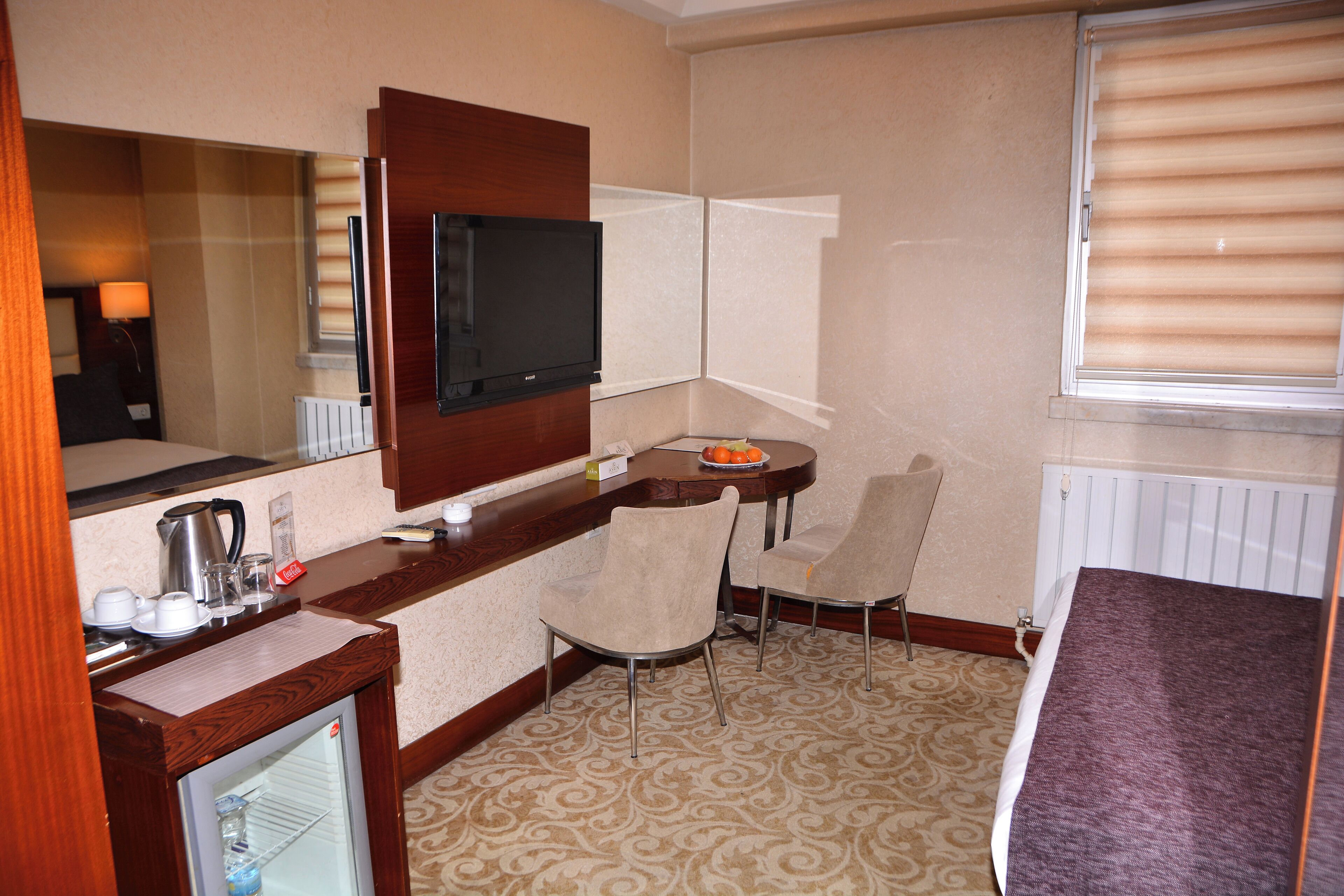 Habitación de hotel con cama, televisión montada en la pared, una pequeña mesa con sillas, un refrigerador compacto y una ventana con persianas.
