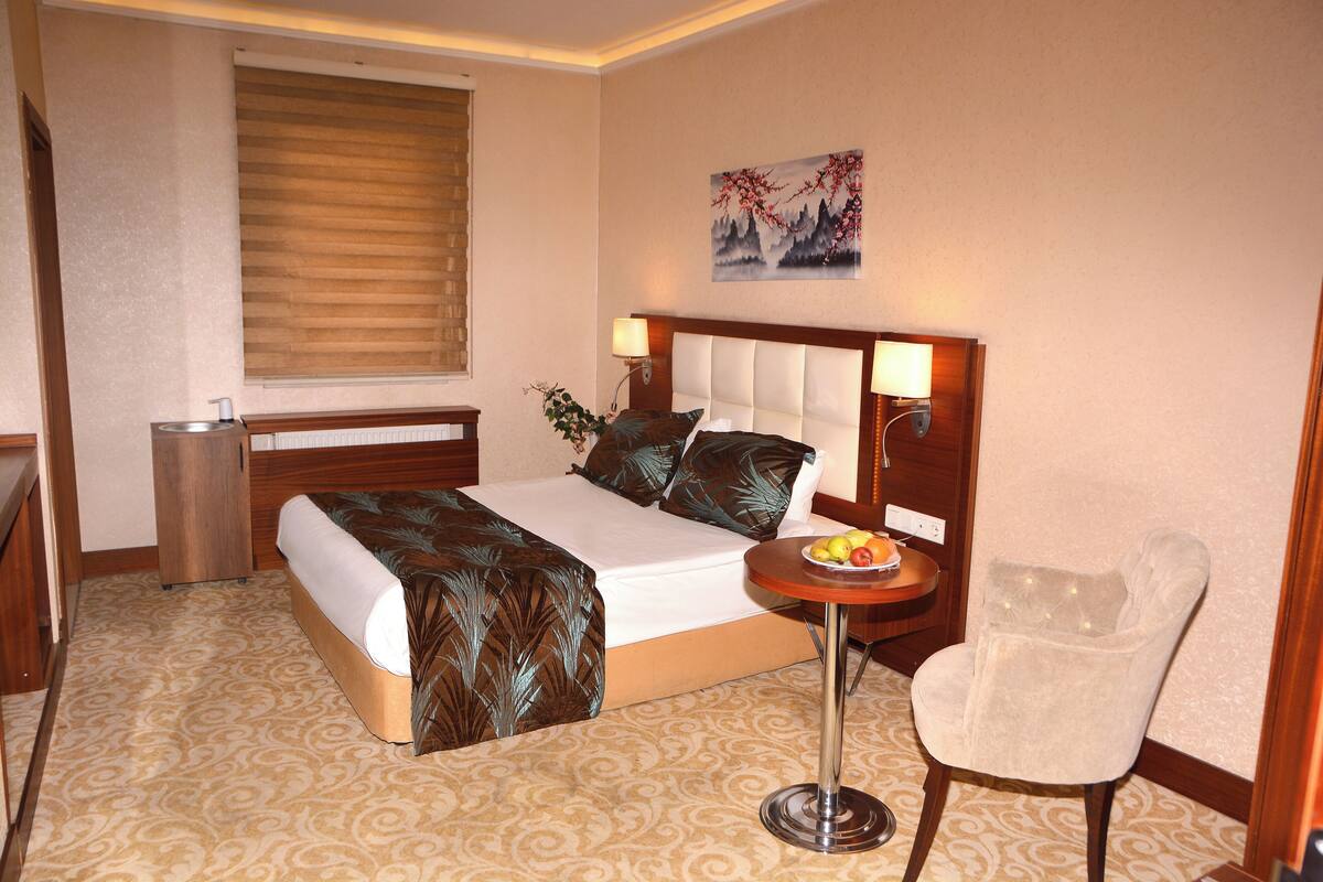 A hotel room with a bed, bedside table with a lamp, a chair, and a small table with fruit.