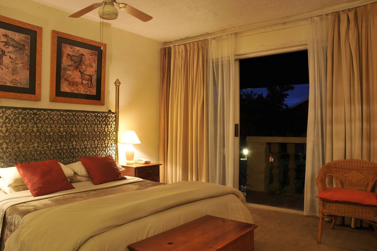 A hotel room with a bed, a nightstand, a lamp, a wicker chair, and a balcony with curtains.