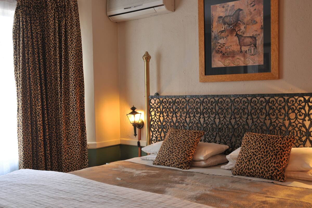 A hotel room with a bed, leopard print curtains, a wall-mounted air conditioner, a framed artwork, and a decorative iron headboard.