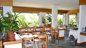 Un comedor amplio con varias mesas y sillas de madera, ventanas grandes con vistas a zonas verdes y plantas en macetas.