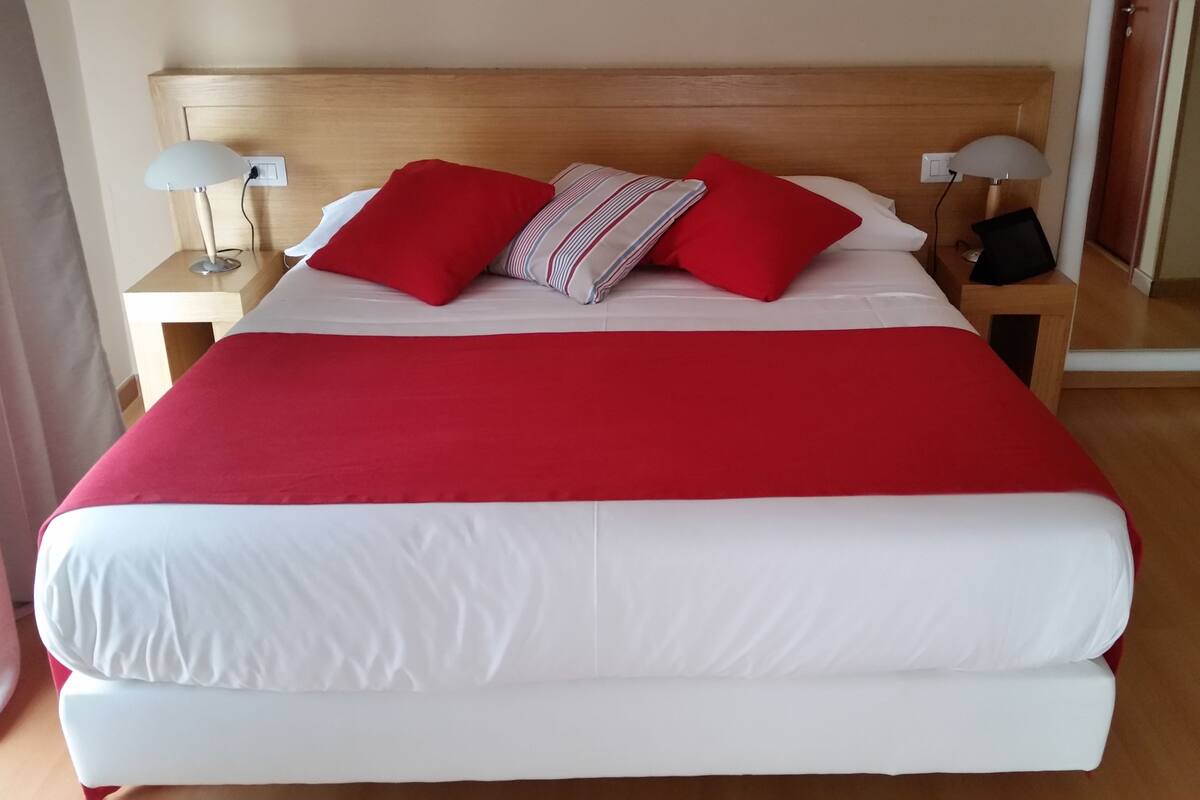 A neatly made bed with a red and white bedspread, two red pillows, and a striped pillow. There are two bedside lamps on wooden nightstands.