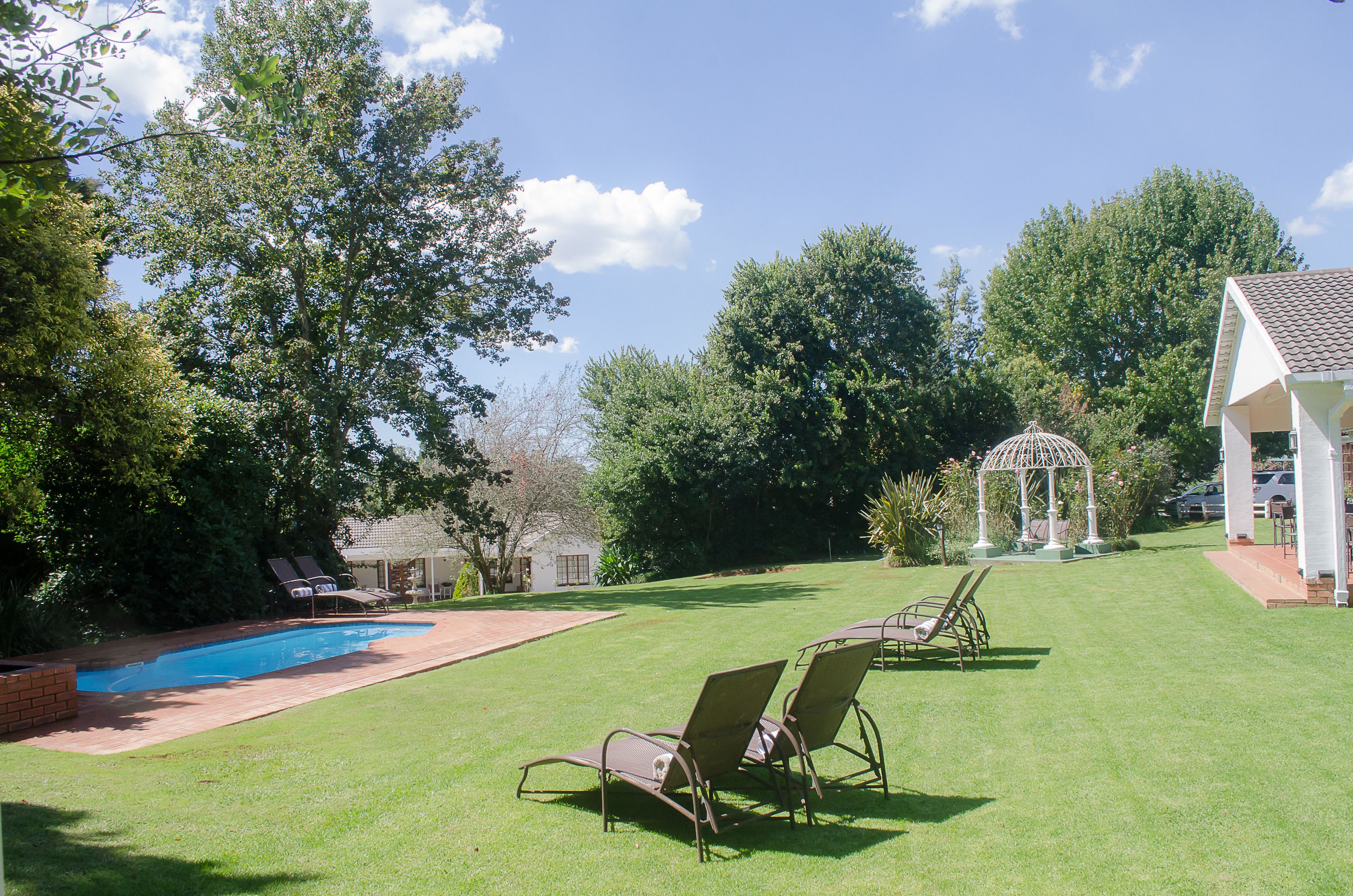 A backyard with a swimming pool, lawn chairs, a gazebo, and a house.