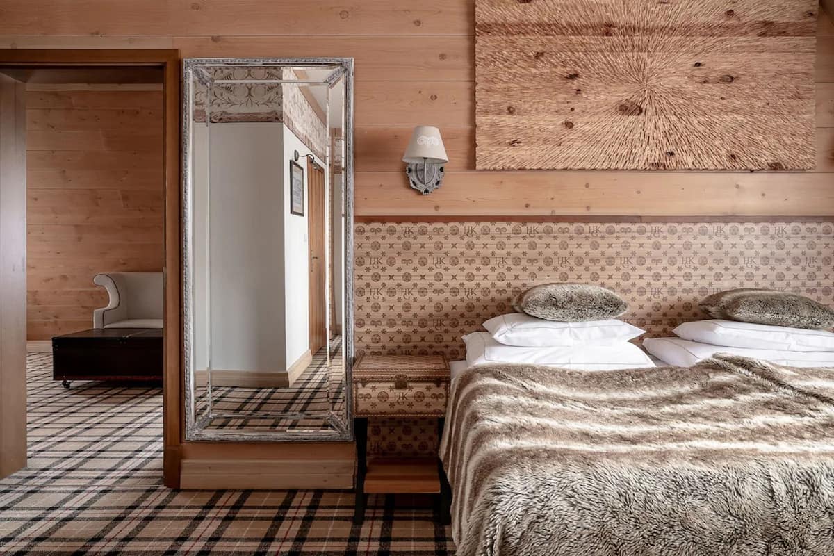 A bedroom with a large mirror, a bed with a fluffy blanket, a bedside table with a lamp, and a patterned wall.