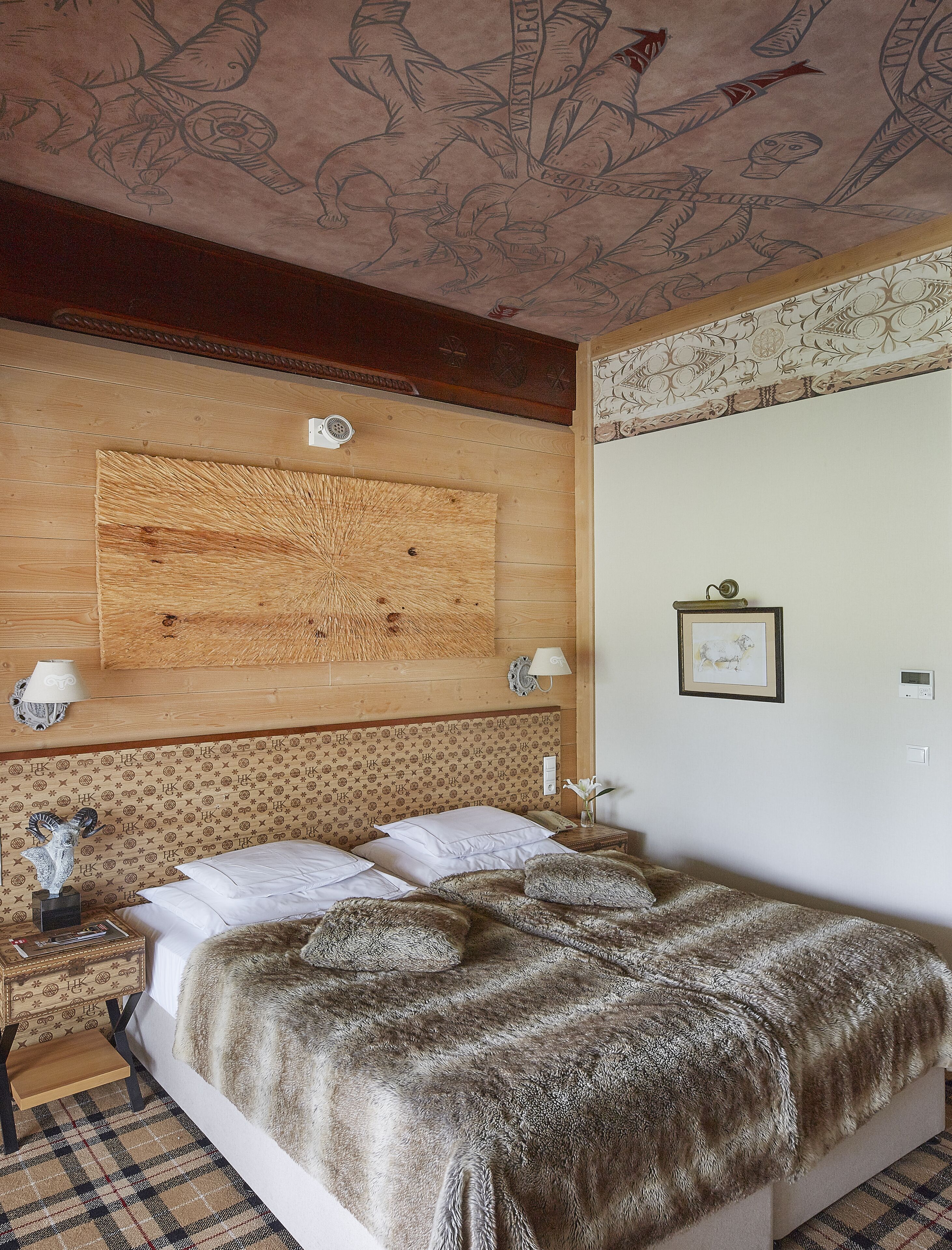 Una habitación de hotel con una cama grande, cabecera de madera, un reloj de pared, un cuadro enmarcado y una mesita de noche con lámpara.