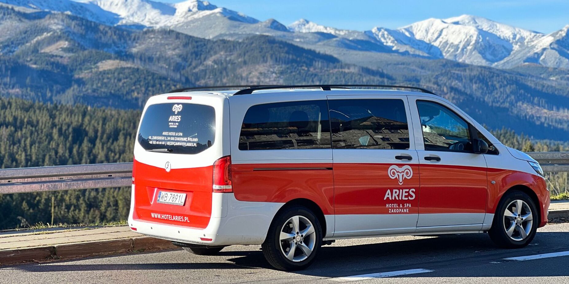 A white and red van with the logo "ARIES HOTEL & SPA ZAKOPANE" on the side, parked on a road with a mountainous backdrop.