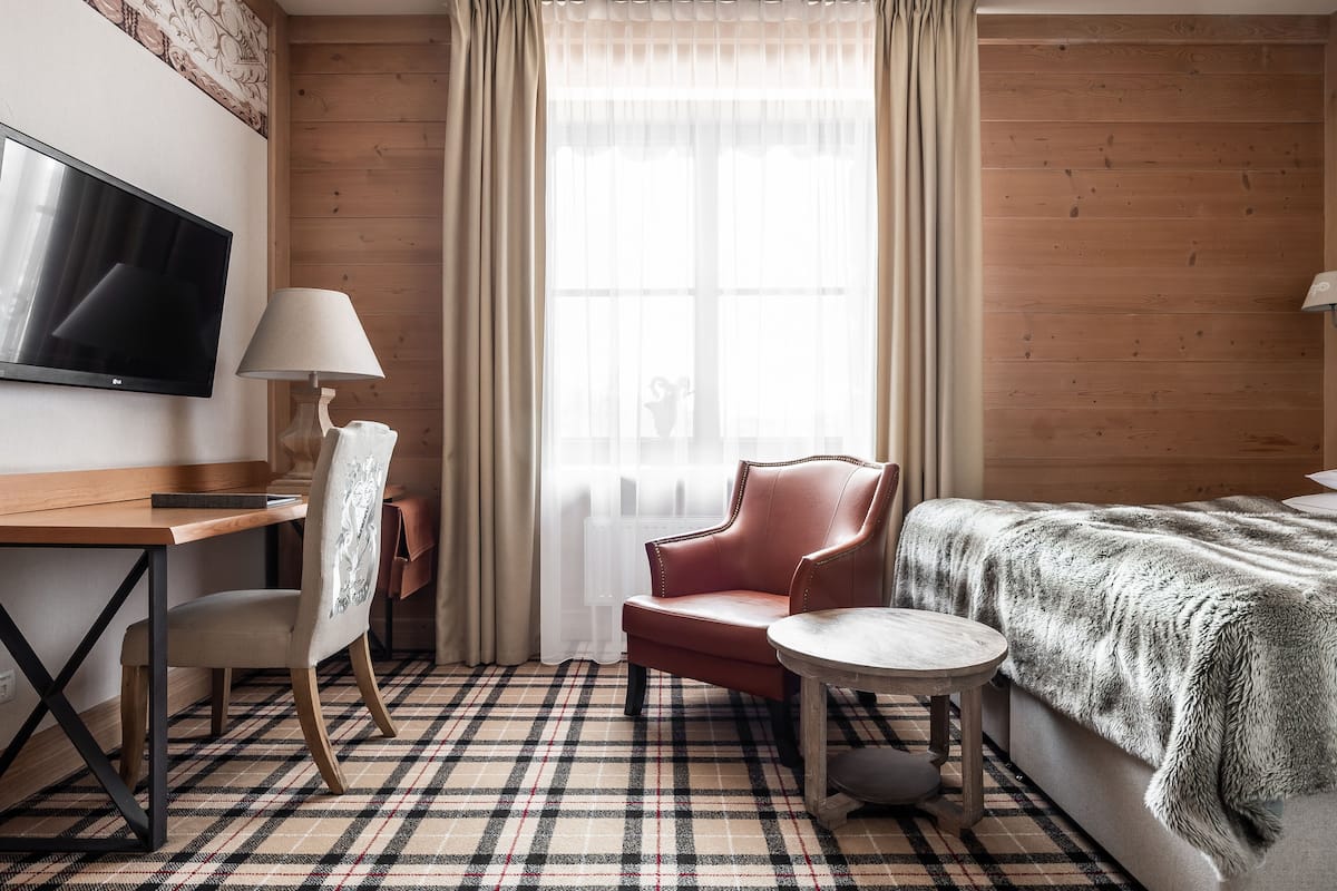 A hotel room with a wooden floor, a bed with a fur throw, a desk with a TV, a chair, and a lamp.