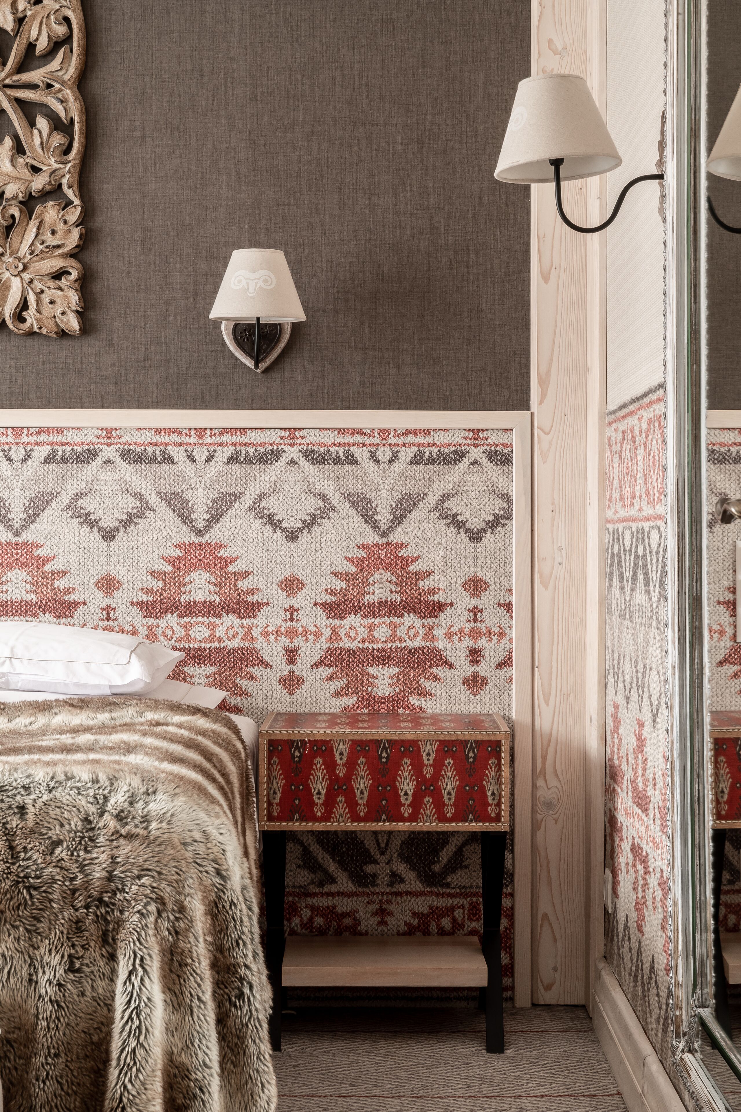 Una habitación con un cabecero estampado, una cama con una manta de piel, un banco con un cojín blanco y rojo estampado, y dos lámparas fijadas a la pared.