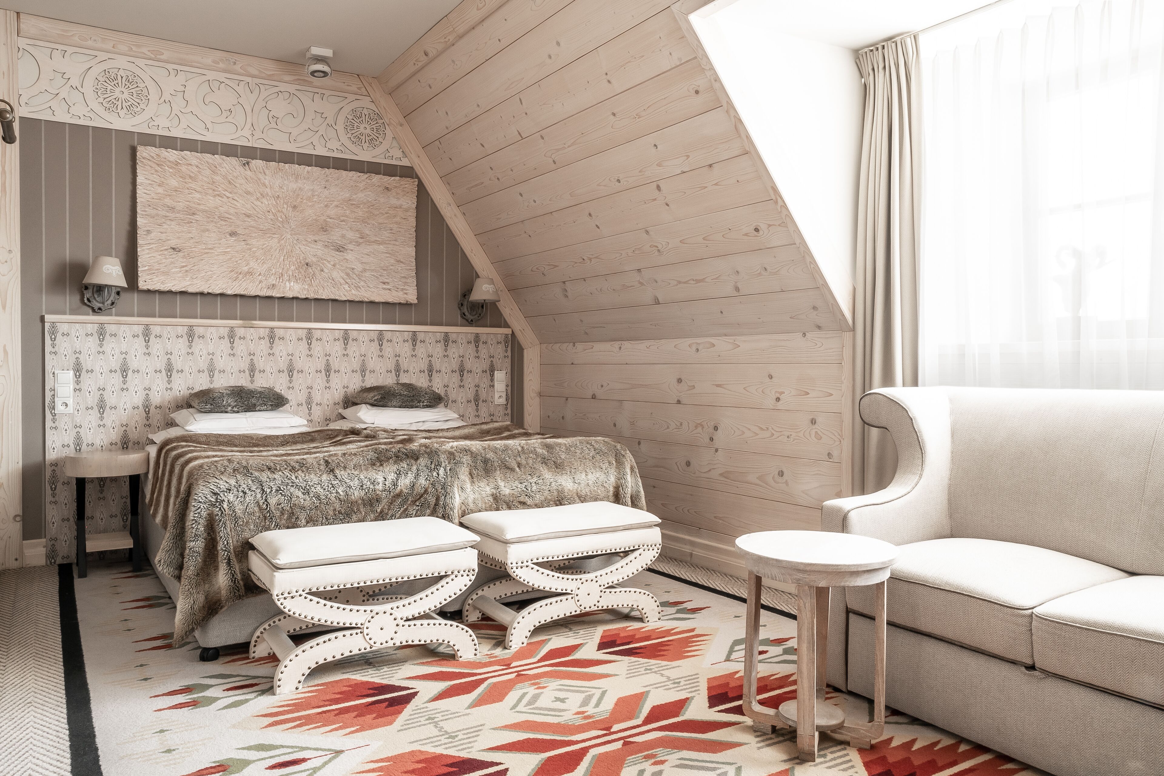 Un dormitorio con techo inclinado, una cama con ropa de cama estampada, dos taburetes blancos tapizados, un sofá blanco y una alfombra con diseño.