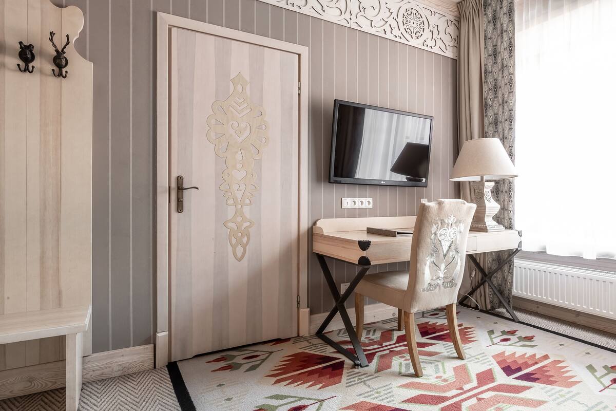 A room with a wooden wardrobe, a desk with a chair, a television, and a patterned rug.