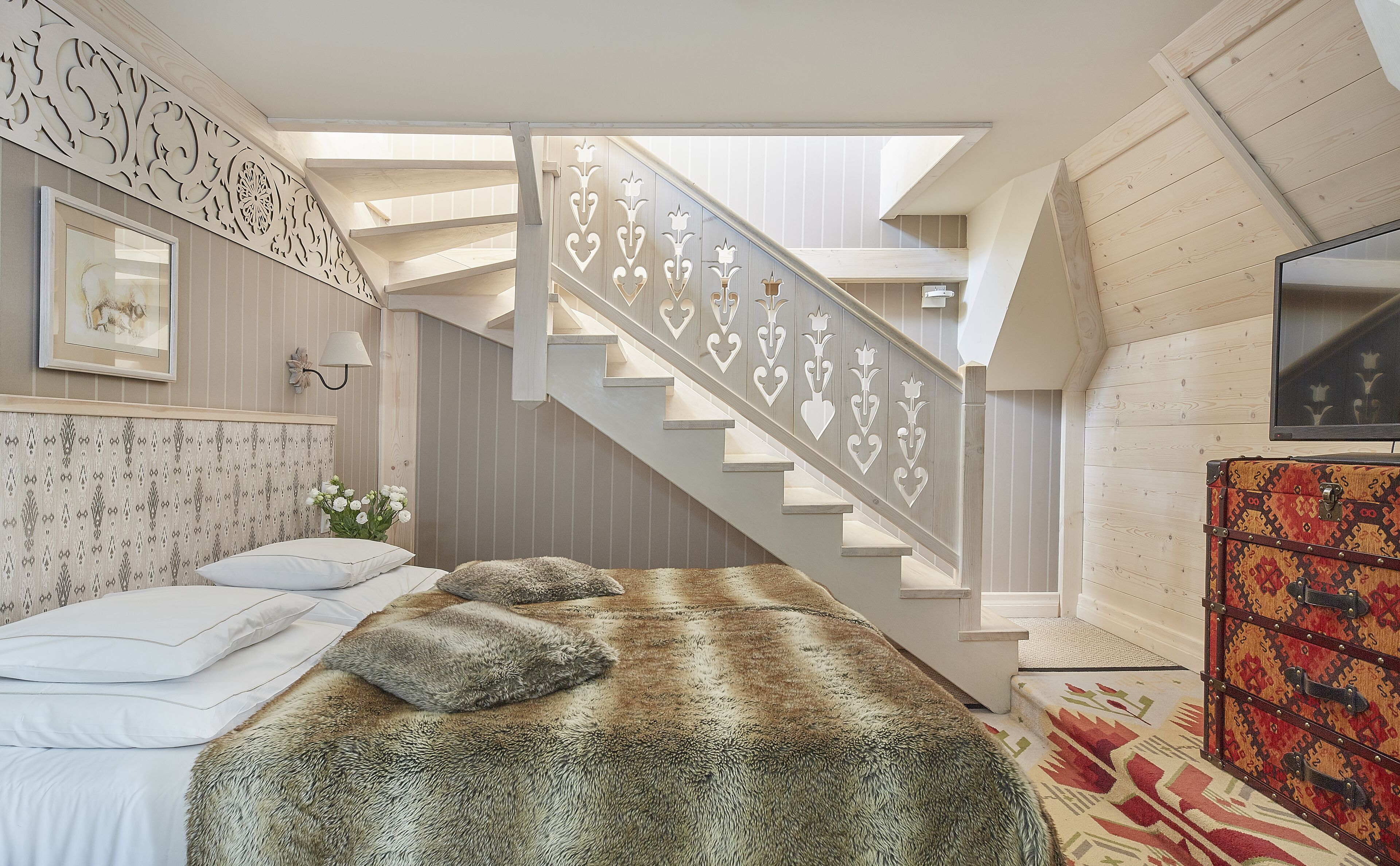 A bedroom with a large bed, a staircase with decorative railings, a television, and a patterned rug.