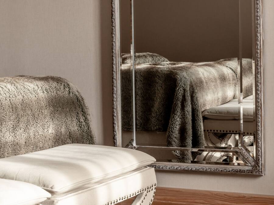 Una habitación con un espejo grande que refleja una cama y un banco blanco tapizado.