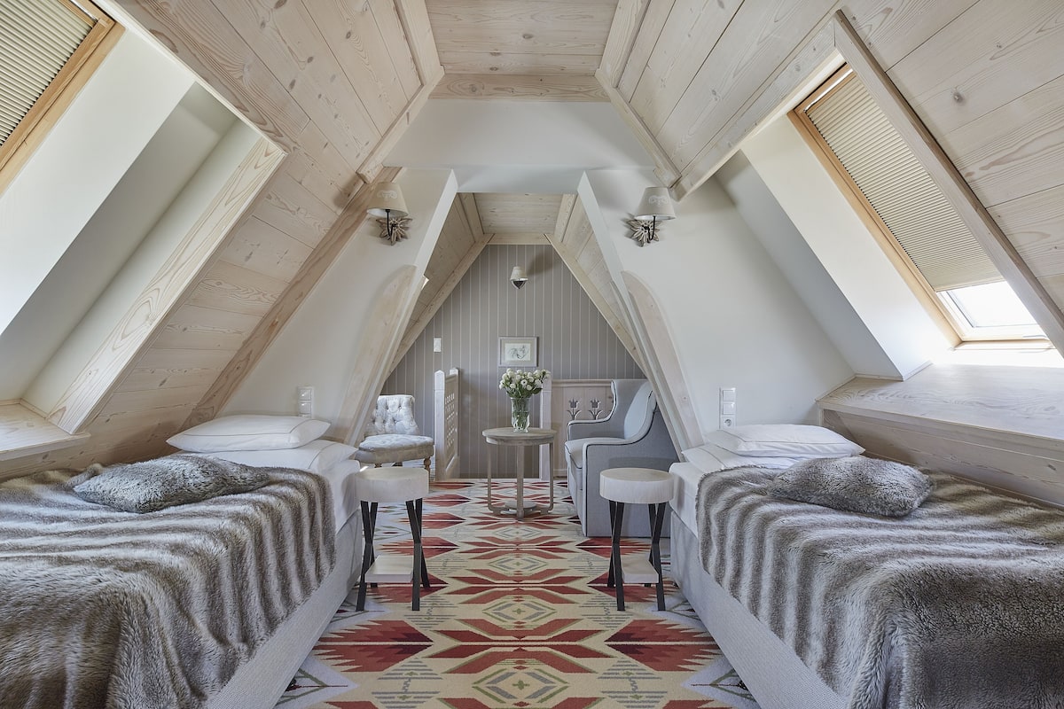 A cozy attic bedroom with a sloped ceiling, two beds with matching headboards, a central table with a vase, and a chair.