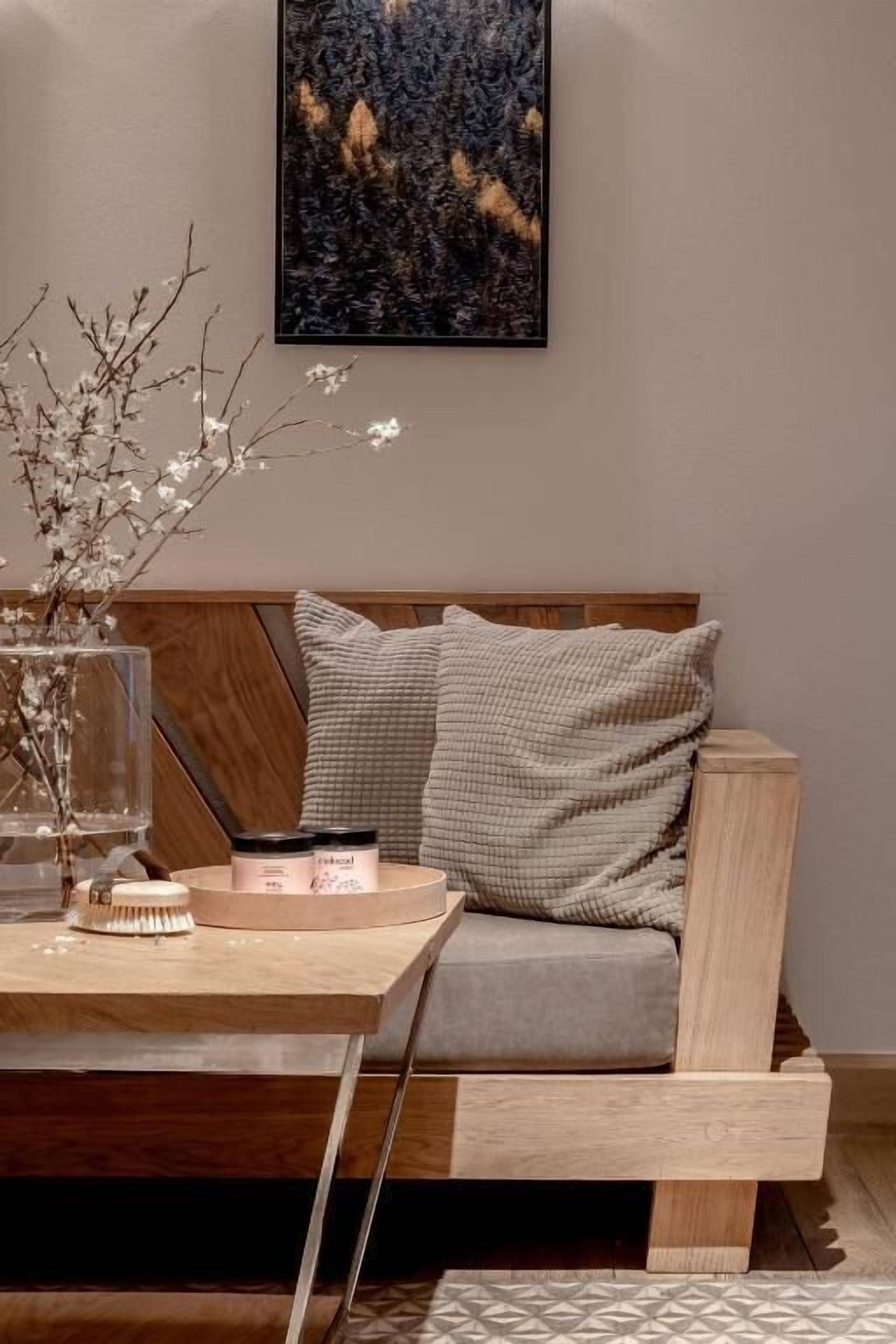 A modern living room with a wooden sofa, a glass vase with branches, and a small table with a candle and a container.