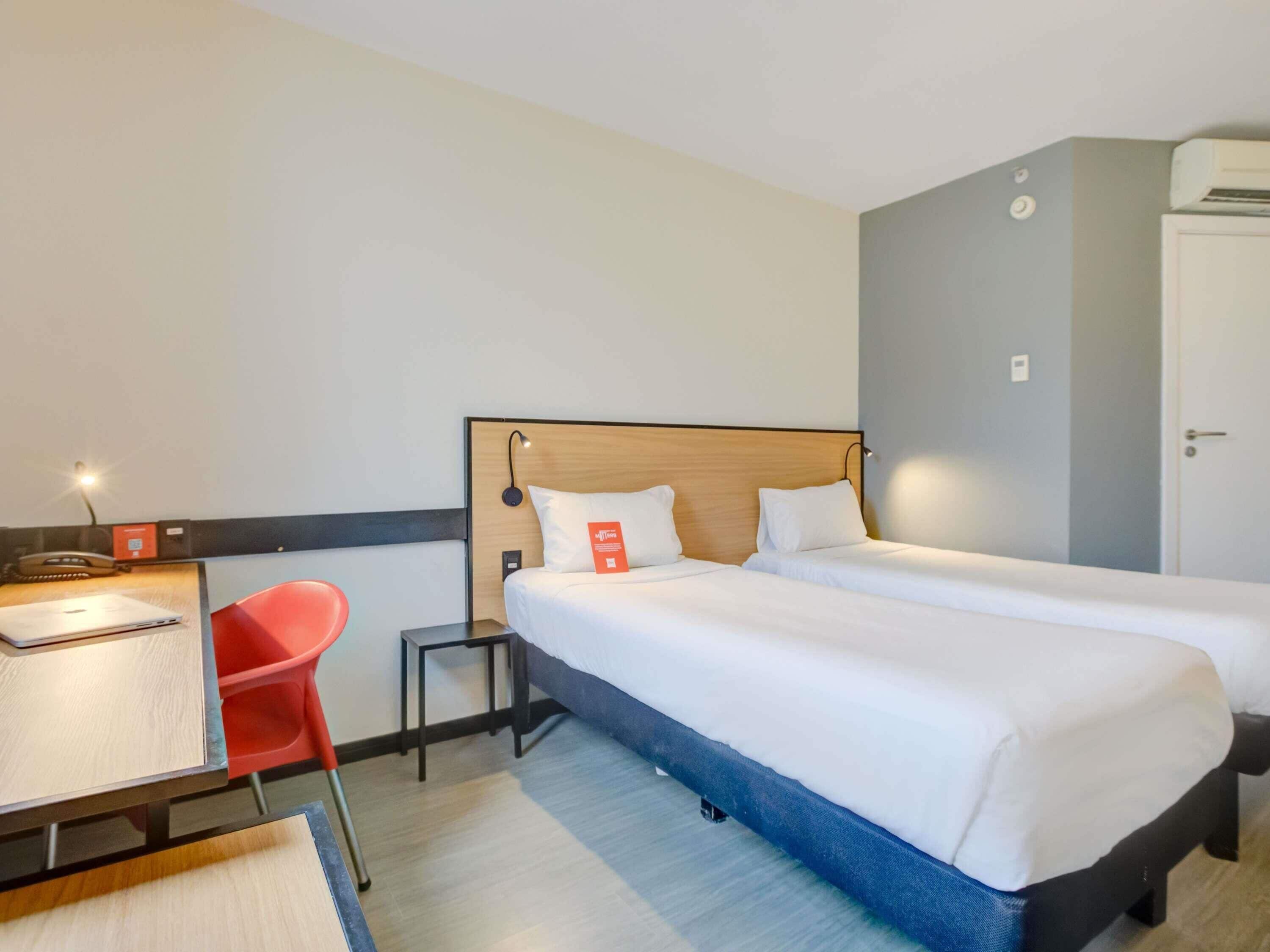 Quarto de hotel com duas camas, uma mesa de escritório com cadeira, uma mesinha e um ar condicionado.