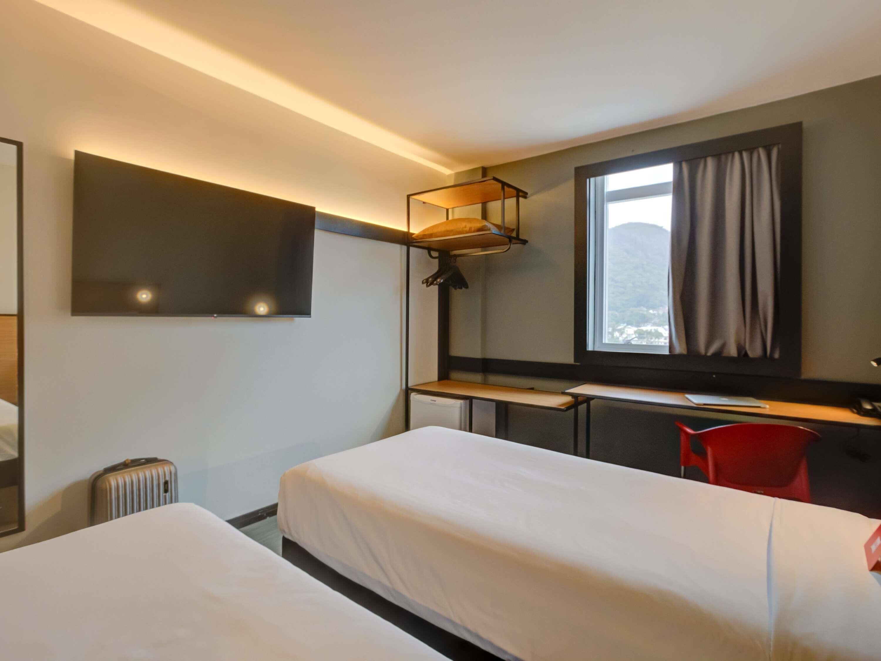 A hotel room with two beds, a desk, a chair, a TV, and a window with curtains.