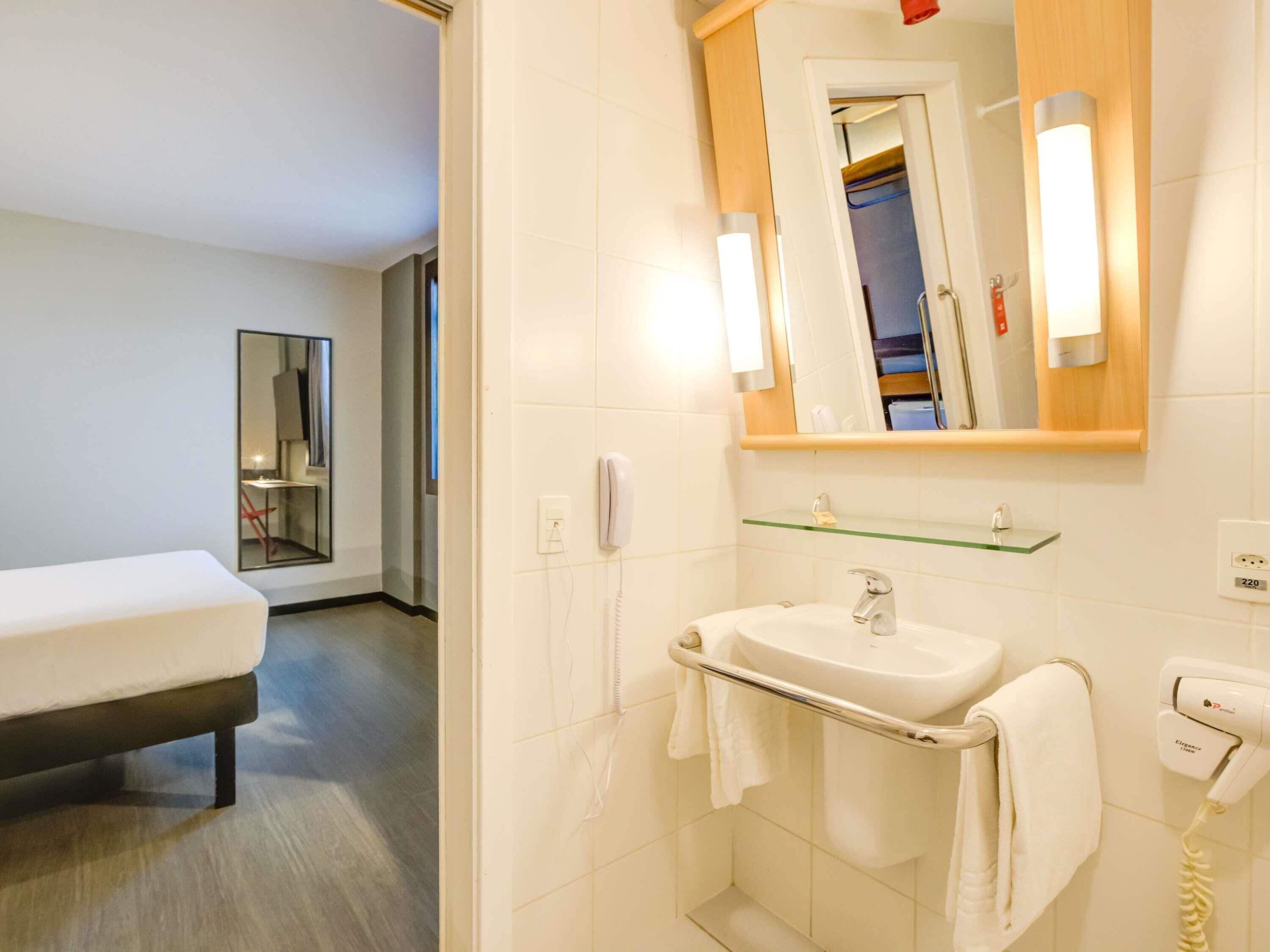 A hotel bathroom with a bed, a sink, a mirror, a towel rack, and a hairdryer.