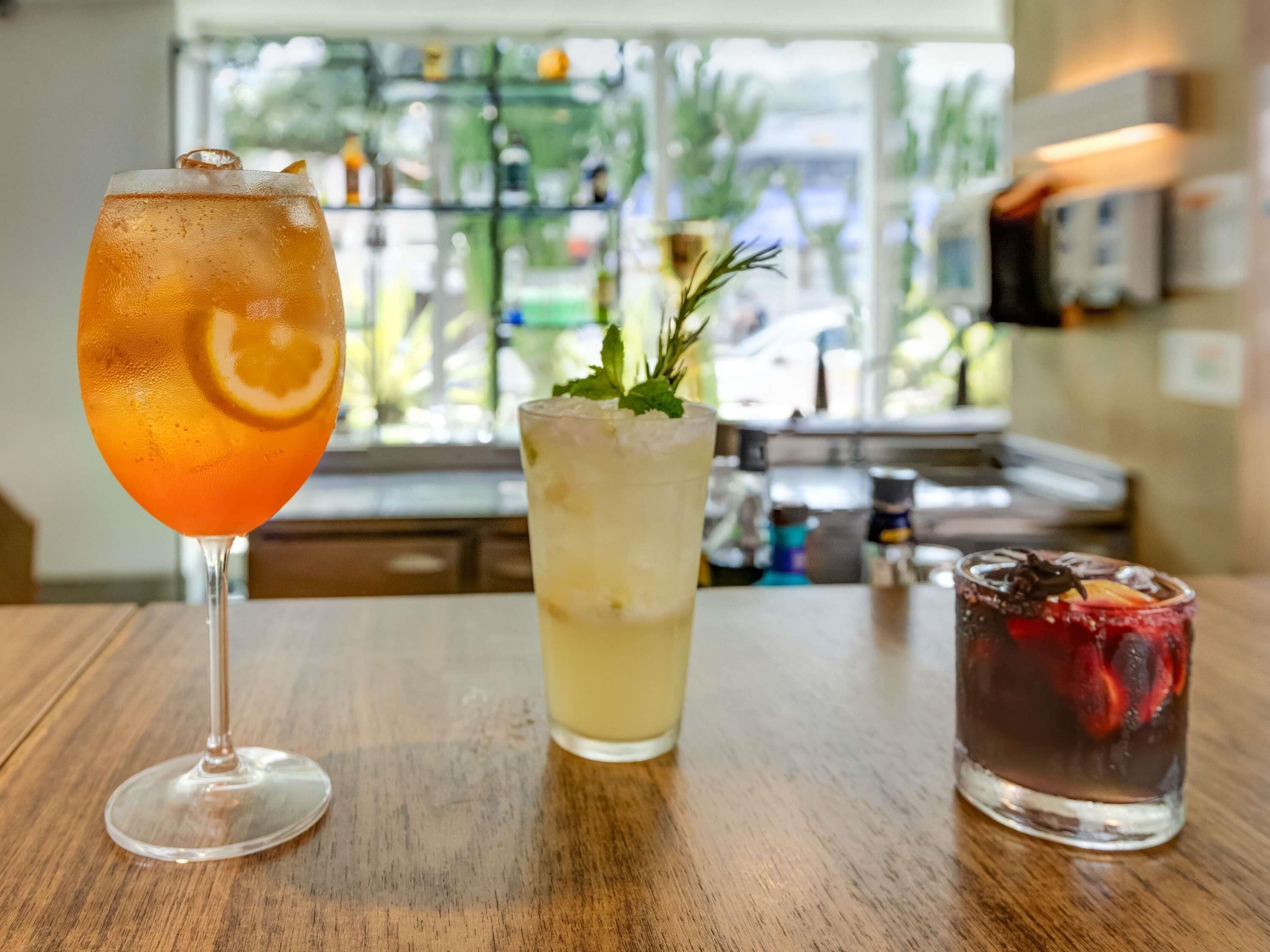 Three drinks on a wooden table with a view of a bar and greenery outside.