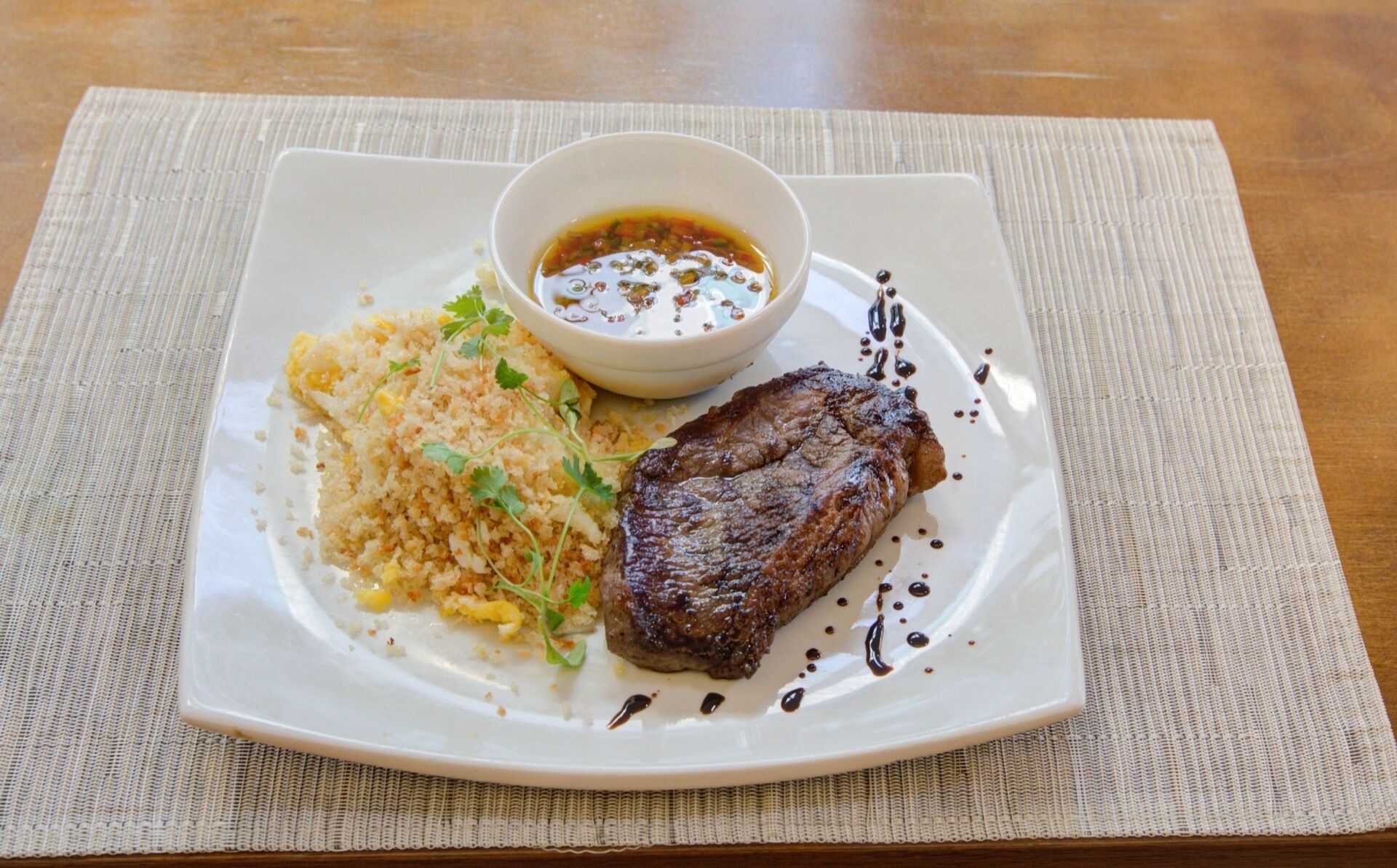 A plate with a steak, rice, and a small bowl of sauce.