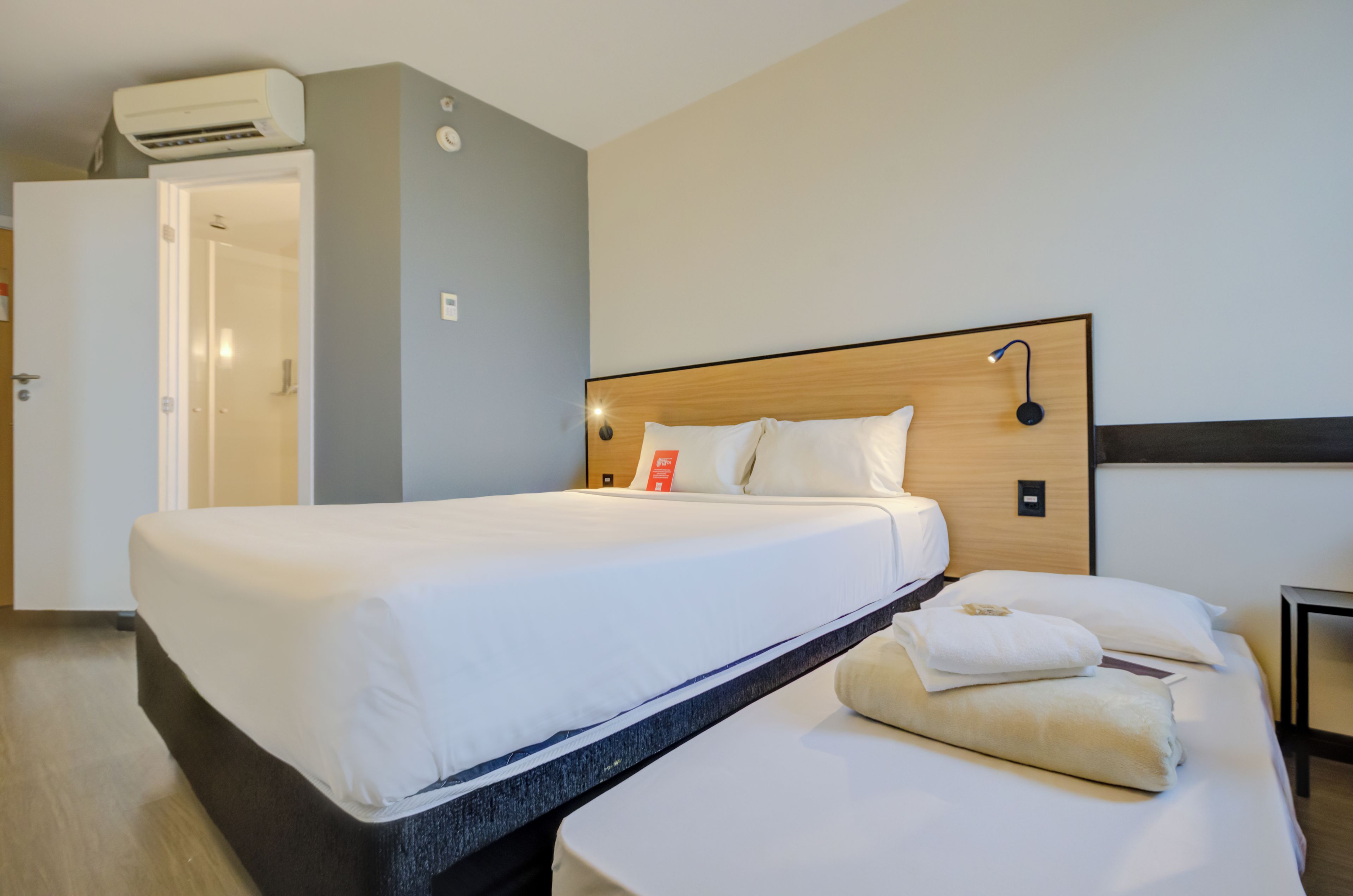 Ein Hotelzimmer mit Bett, Nachttisch, Klimaanlage und einer Tür zu einem weiteren Raum.