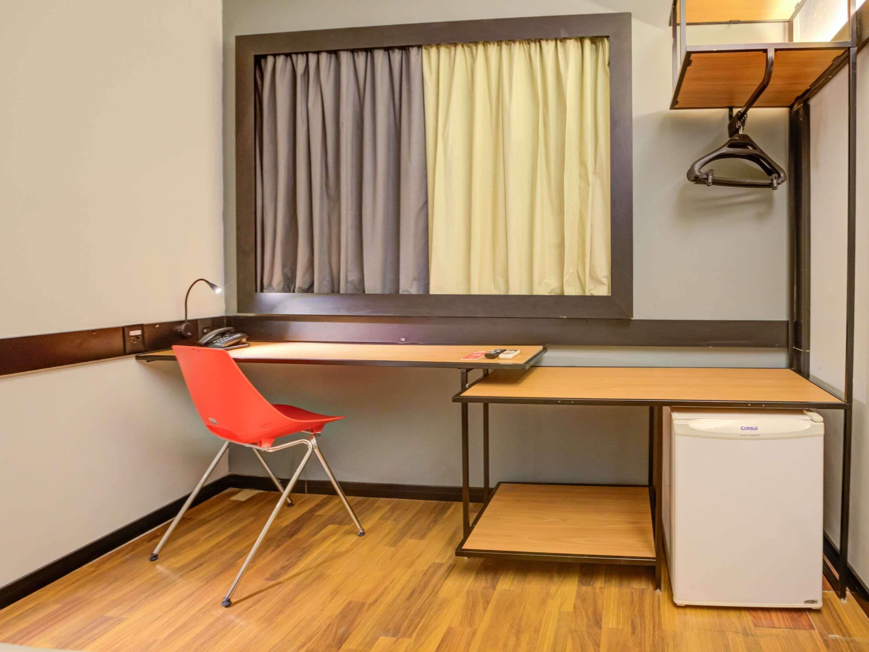 Una habitación compacta con escritorio de madera, una silla roja, un refrigerador blanco pequeño y una ventana con dos cortinas.